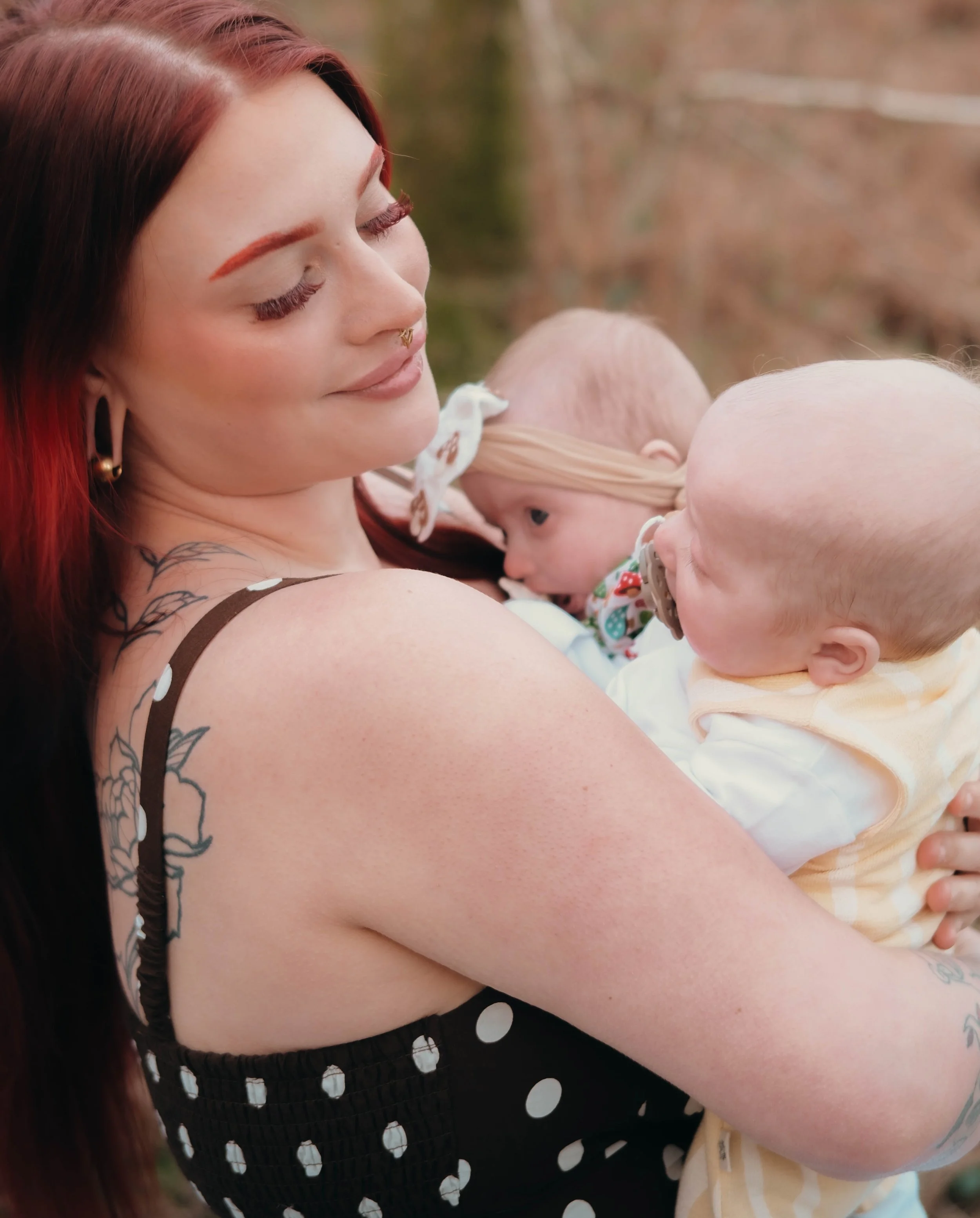 Close-up of a woman with long red hair holding two babies outdoors. The woman has closed eyes and a gentle smile.