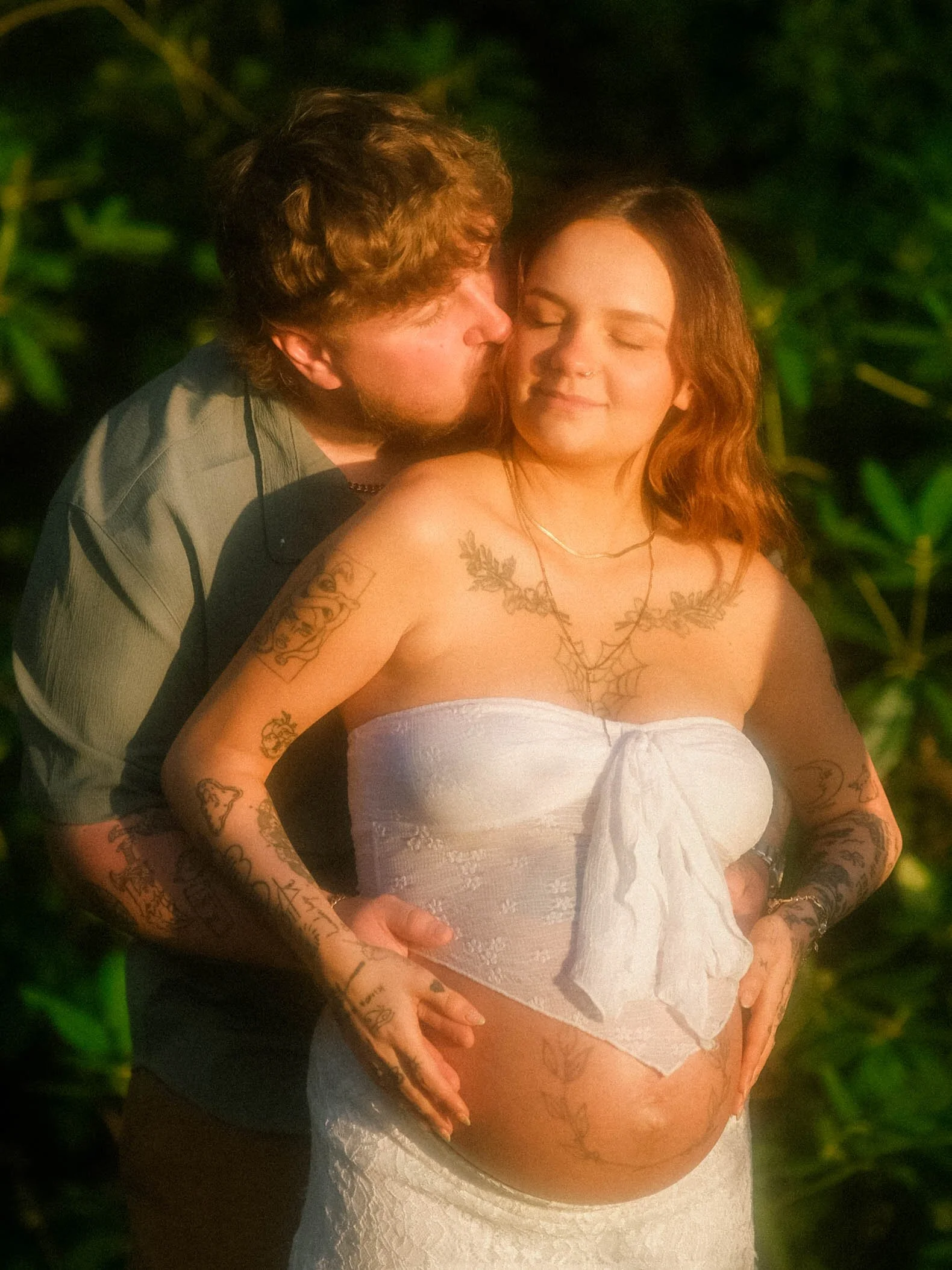 A man and a pregnant woman stand close together outdoors at night, with the man kissing her cheek and her smiling with eyes closed. The woman is wearing a white towel dress and has tattoos on her arms and chest. The man is dressed in dark clothing.