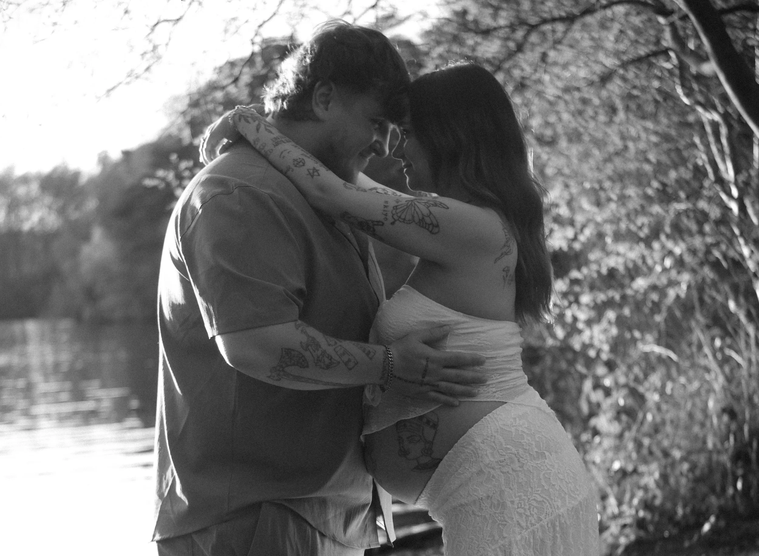 A black and white photo of a couple embracing outdoors with trees in the background, the woman wearing a white dress and the man wearing a jacket, their foreheads touching and eyes closed.