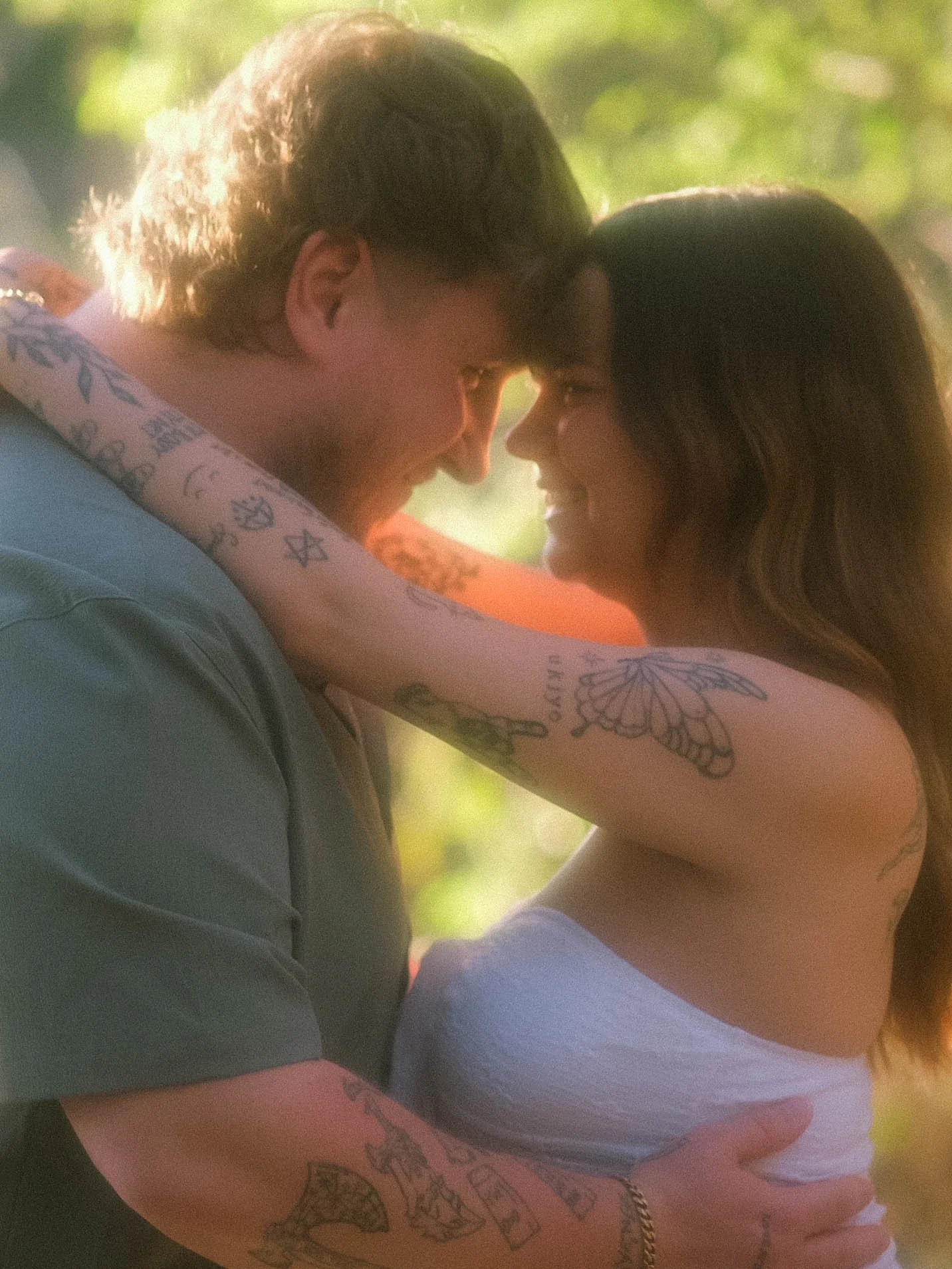 A couple close together outside, touching foreheads and smiling, with a blurred green background.