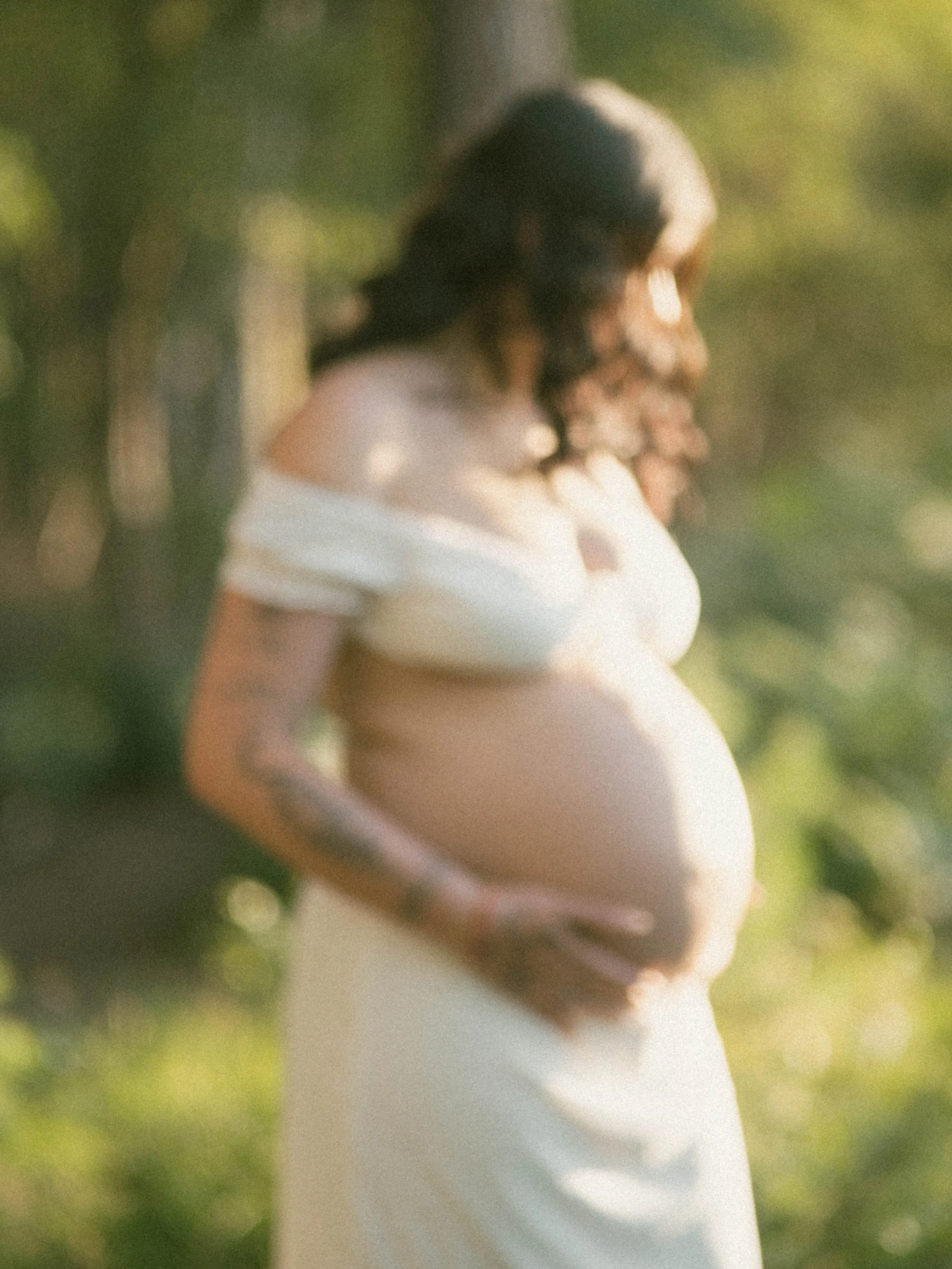 Blurred image of a pregnant woman in a white dress outdoors with greenery in the background.