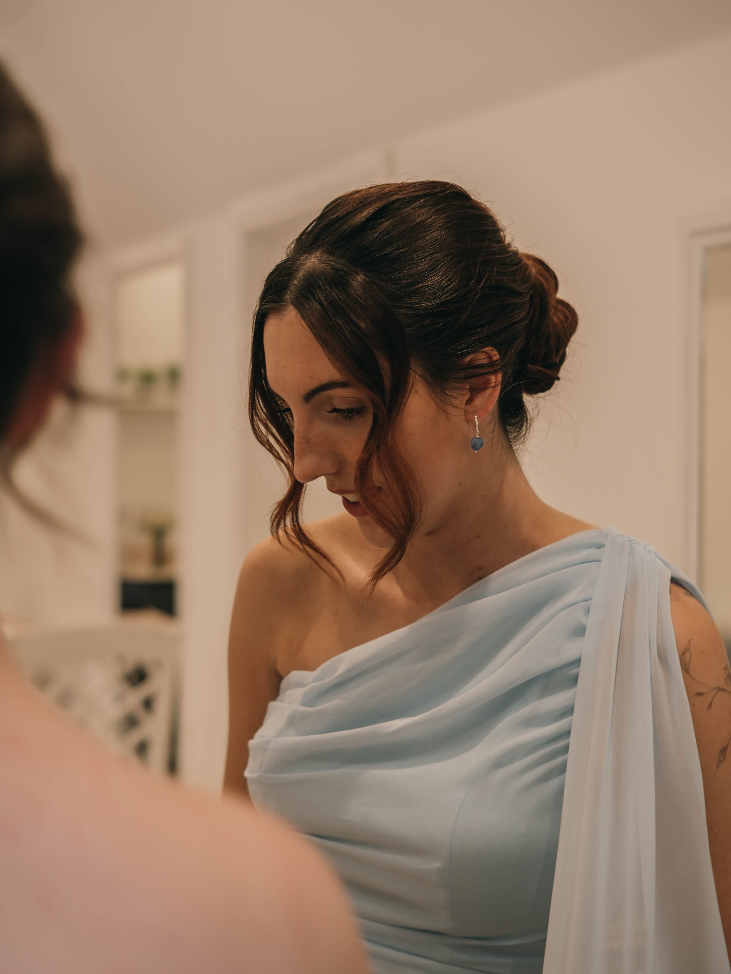A woman with short, dark red hair styled in soft waves, wearing a pearl earring with a blue gemstone, and a white one-shoulder dress, looking down, in a softly lit room.