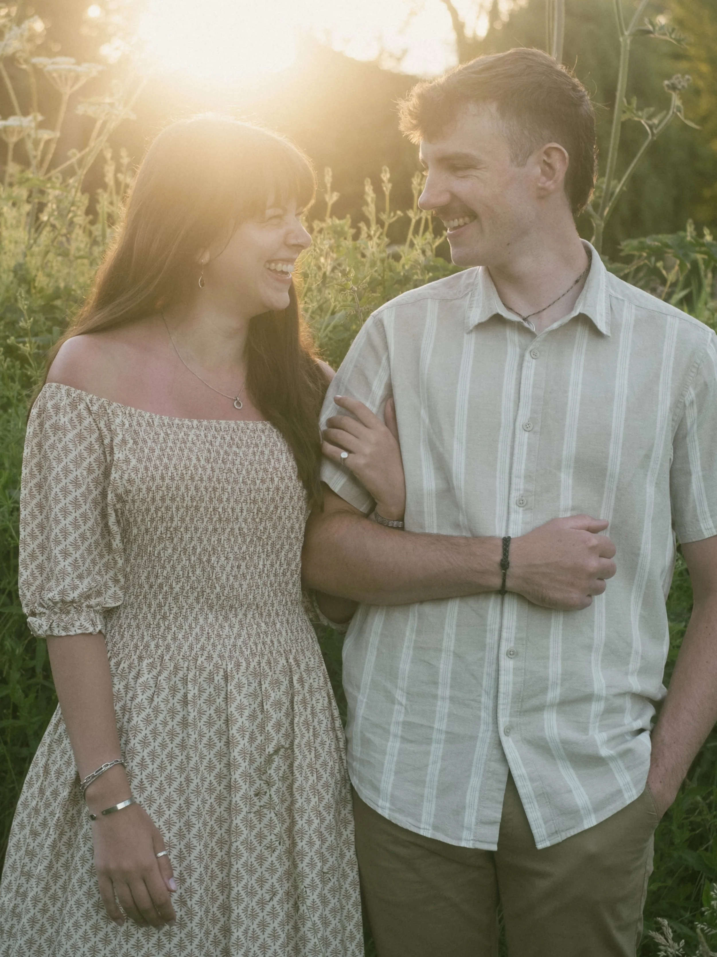 A man and a woman smiling and looking at each other outdoors during sunset, standing in a field with green plants.