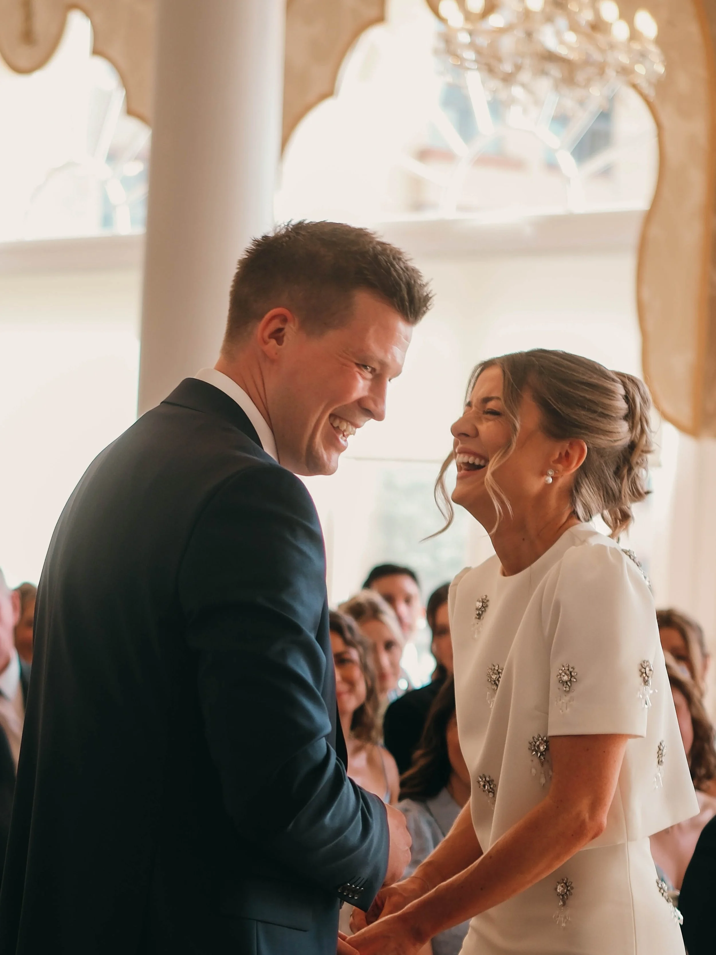 A couple, a groom in a black suit and a bride in a white dress with floral embellishments, are holding hands and smiling during their wedding ceremony, with guests seated in the background.