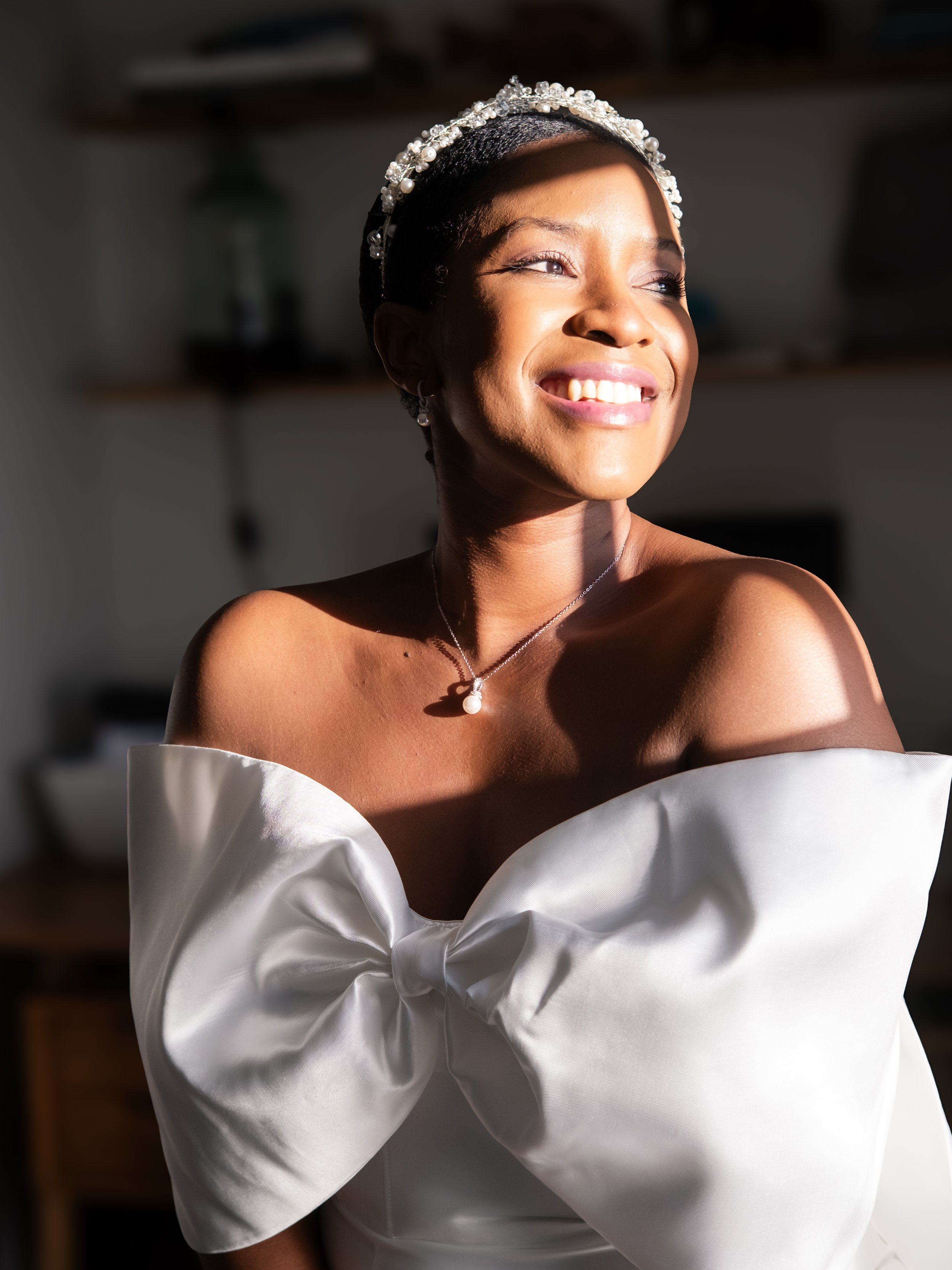 Close-up of a smiling young woman wearing an off-shoulder white gown and a pearl necklace, with a pearl and rhinestone headband, in a brightly lit room.