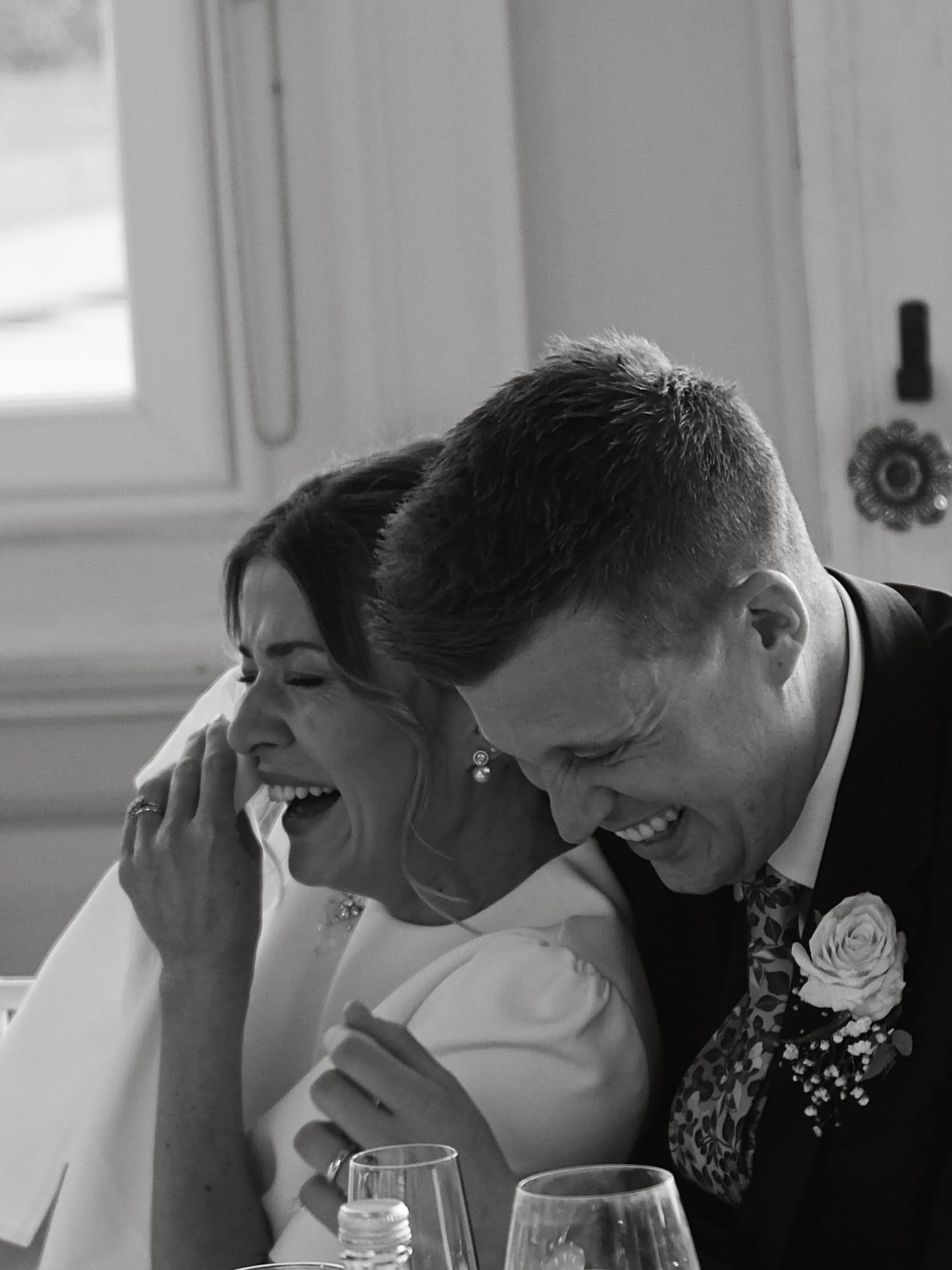 A black and white photo of two people, a man and a woman, sharing a joyful moment at a formal event, possibly a wedding, sitting at a table with wine glasses, a water bottle, and a rose boutonniere.