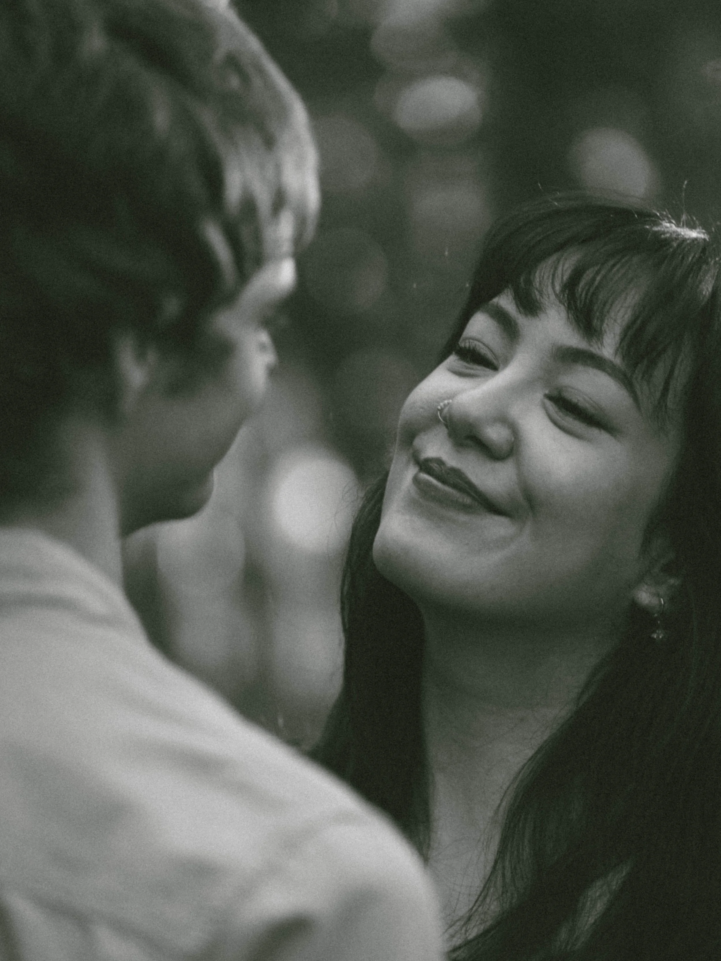 A black-and-white close-up photo of a woman with short hair and a nose ring smiling, looking at a man who is only partially visible in the foreground.