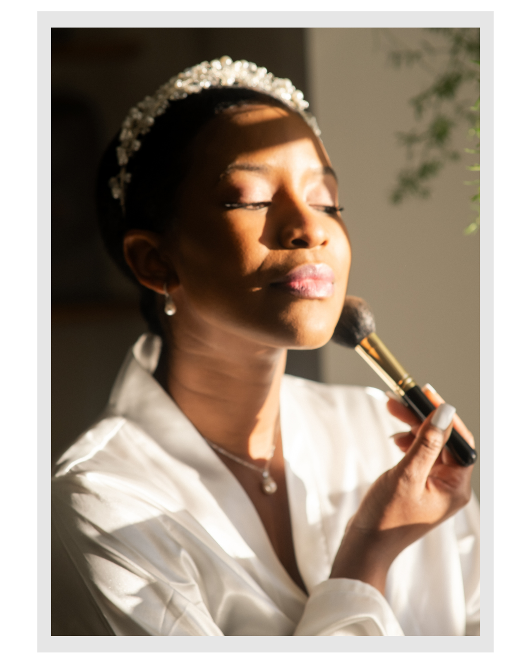 A woman with a tiara and earrings applying makeup with a brush, her eyes closed in a well-lit room.