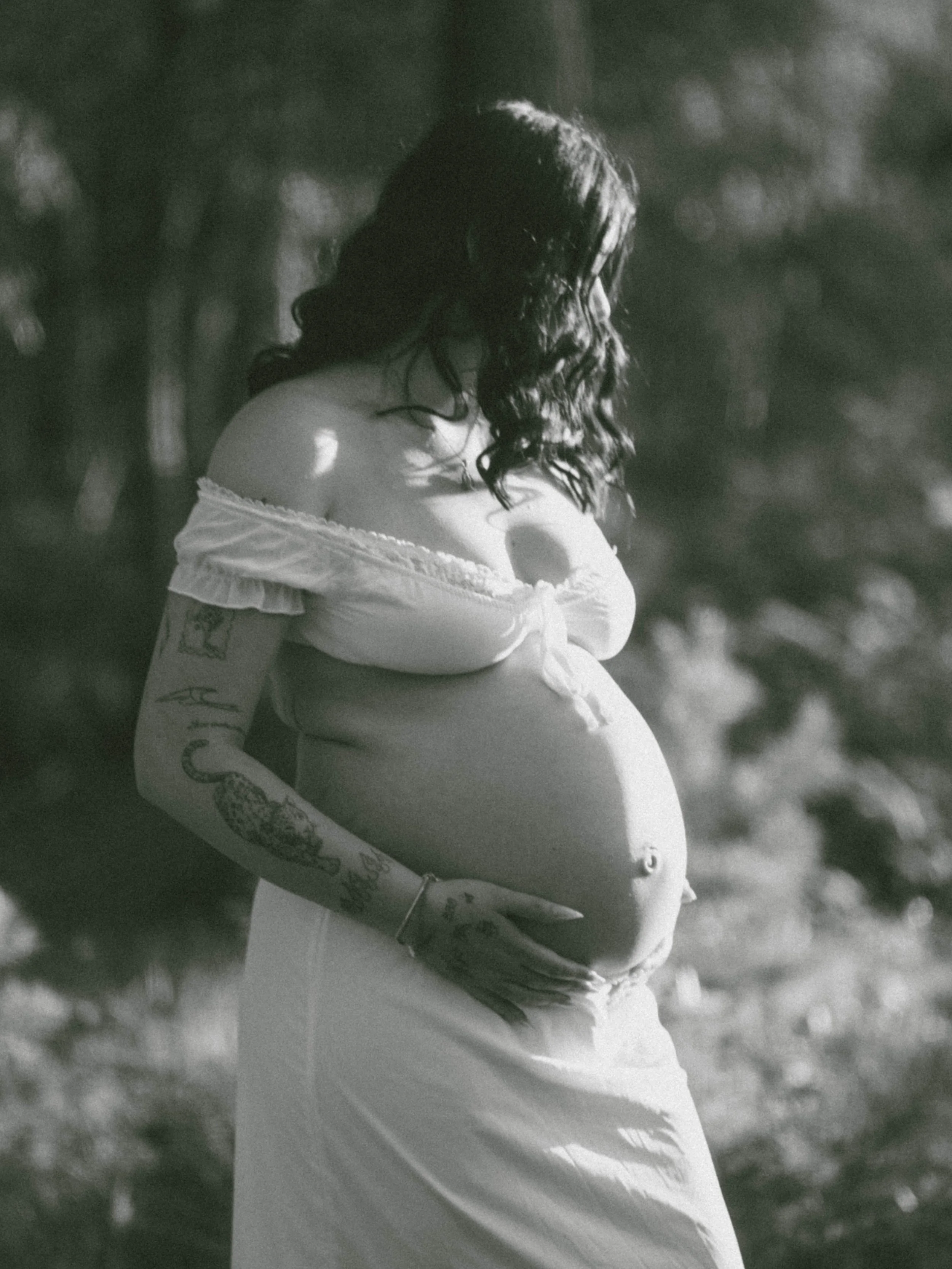 Black and white photo of a pregnant woman with curly hair, wearing a loose off-shoulder top, standing outdoors in a natural setting.