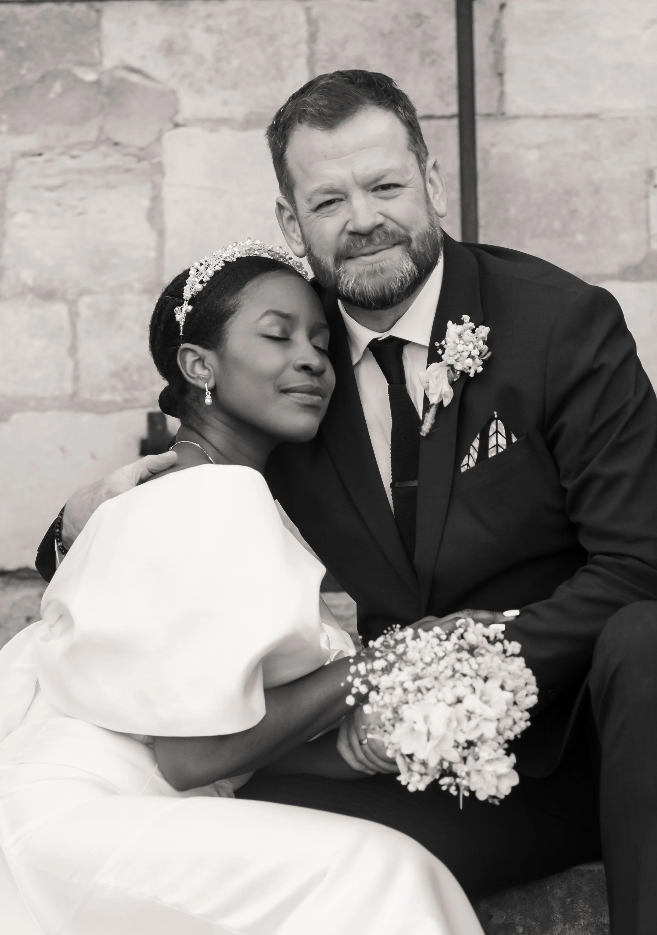 A black and white photo of a bride and groom hugging. The bride has her eyes closed and wears a tiara, earrings, and a white dress. The groom is smiling, has a beard, and wears a black suit with a boutonniere. They are sitting close together, with th
