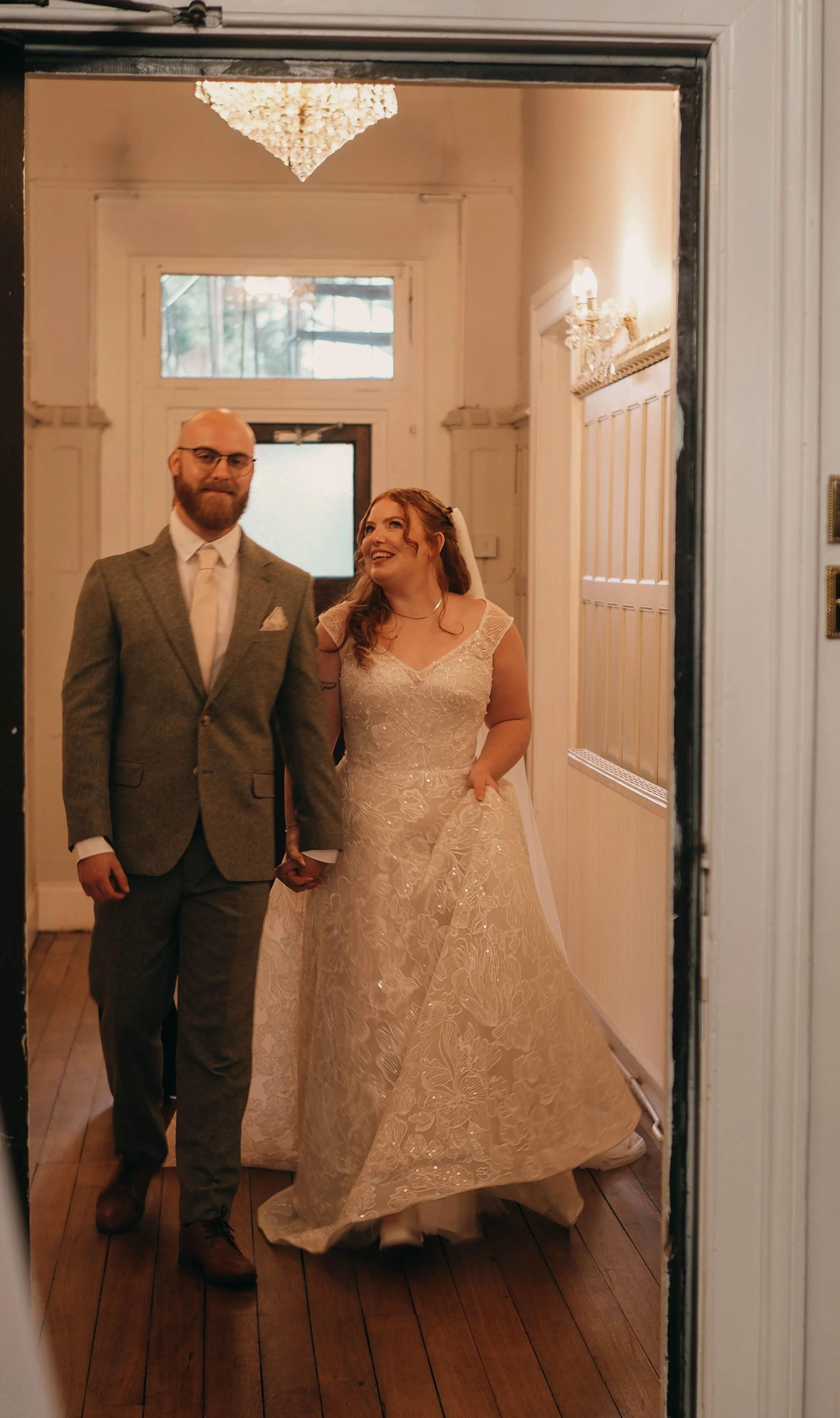 A bride and groom holding hands, taking a mirror selfie at their wedding. The bride is in a cream lace wedding gown, and the groom is in a gray suit with a white shirt and tie. The room behind them has a chandelier and soft lighting.