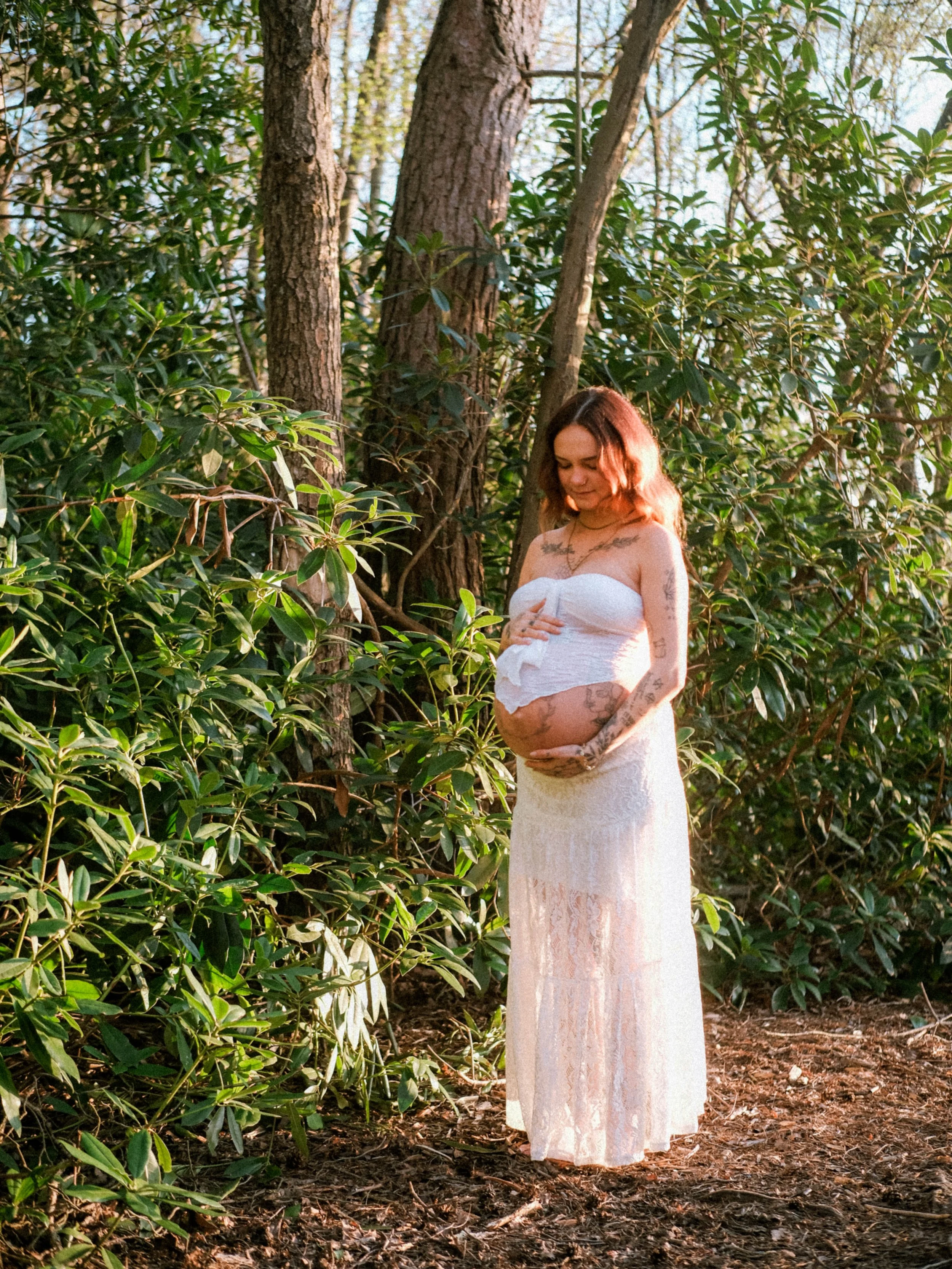 A pregnant woman with tattoos on her arms and chest stands outdoors in a forested area with tall trees and green foliage, wearing a white lace dress and holding her pregnant belly.