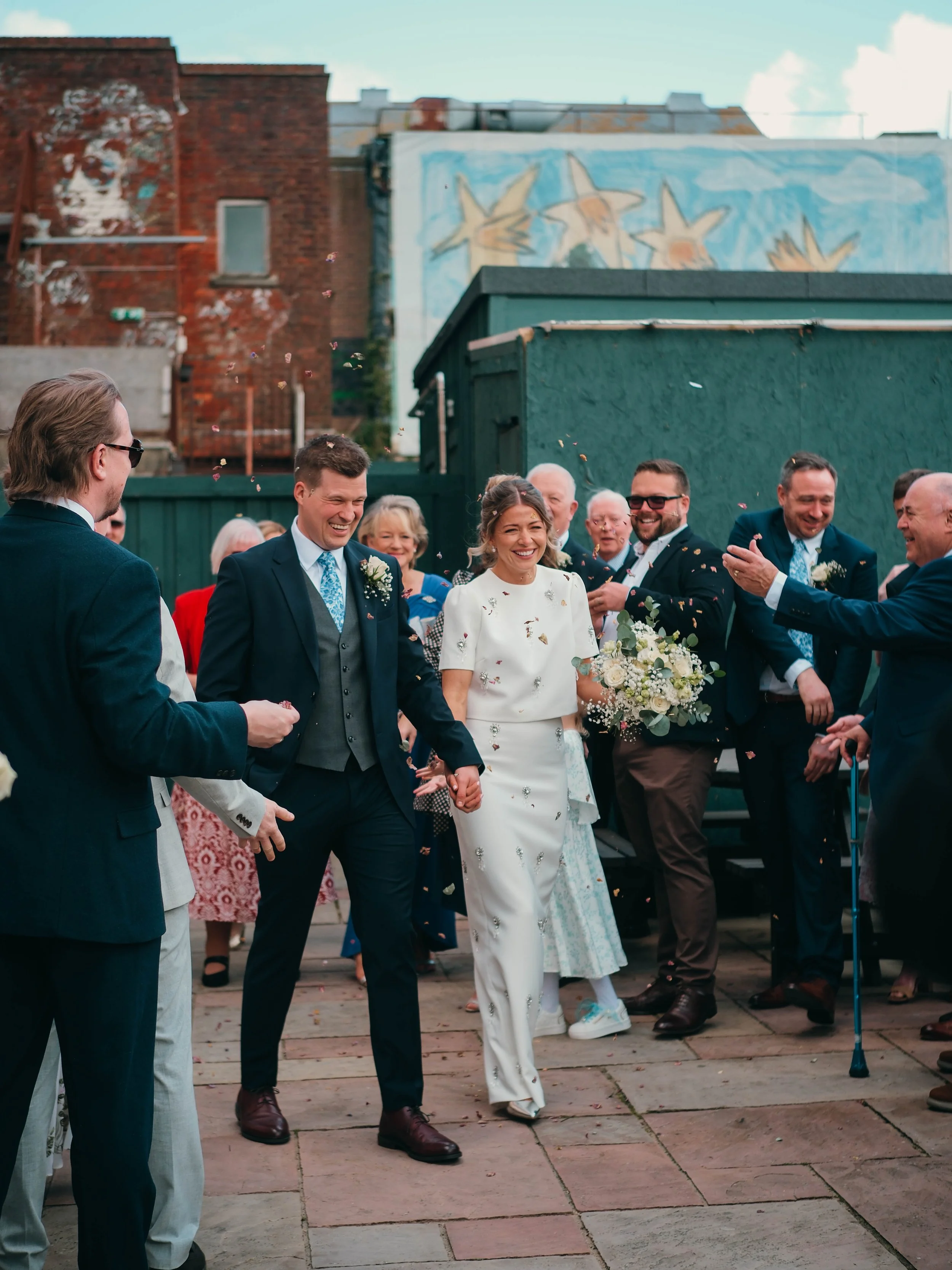 People celebrating at a wedding outside, with a woman in a white wedding dress holding a bouquet, surrounded by friends and family, with confetti falling down.