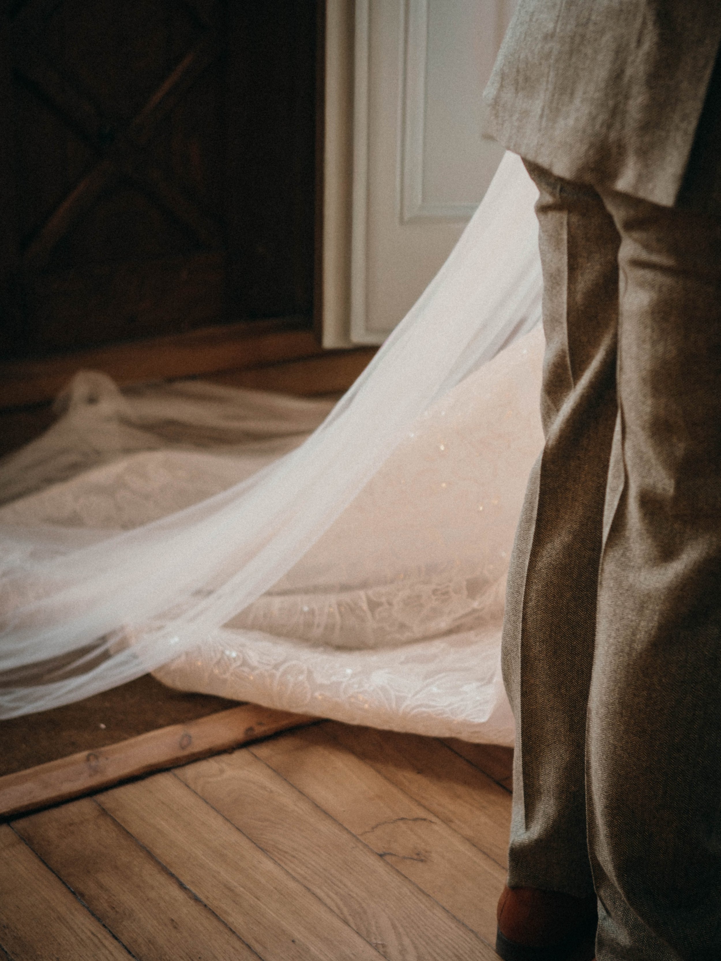 Part of a person wearing gray pants and brown shoes standing on a wooden floor next to a white lace curtain and part of a white door in the background.
