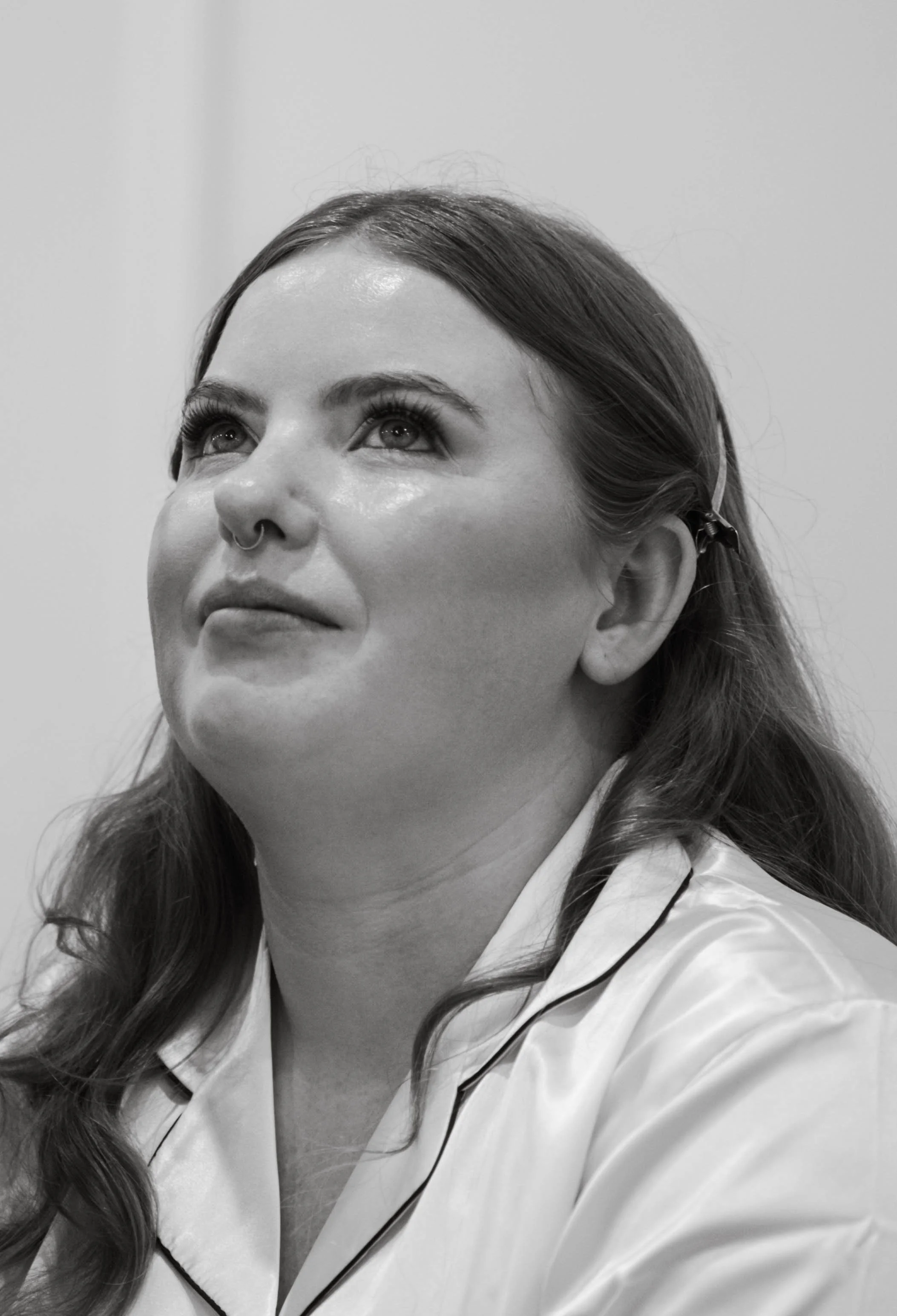 Close-up black-and-white photo of a woman with long hair, looking up and to her right, wearing a white blouse and a nose ring.