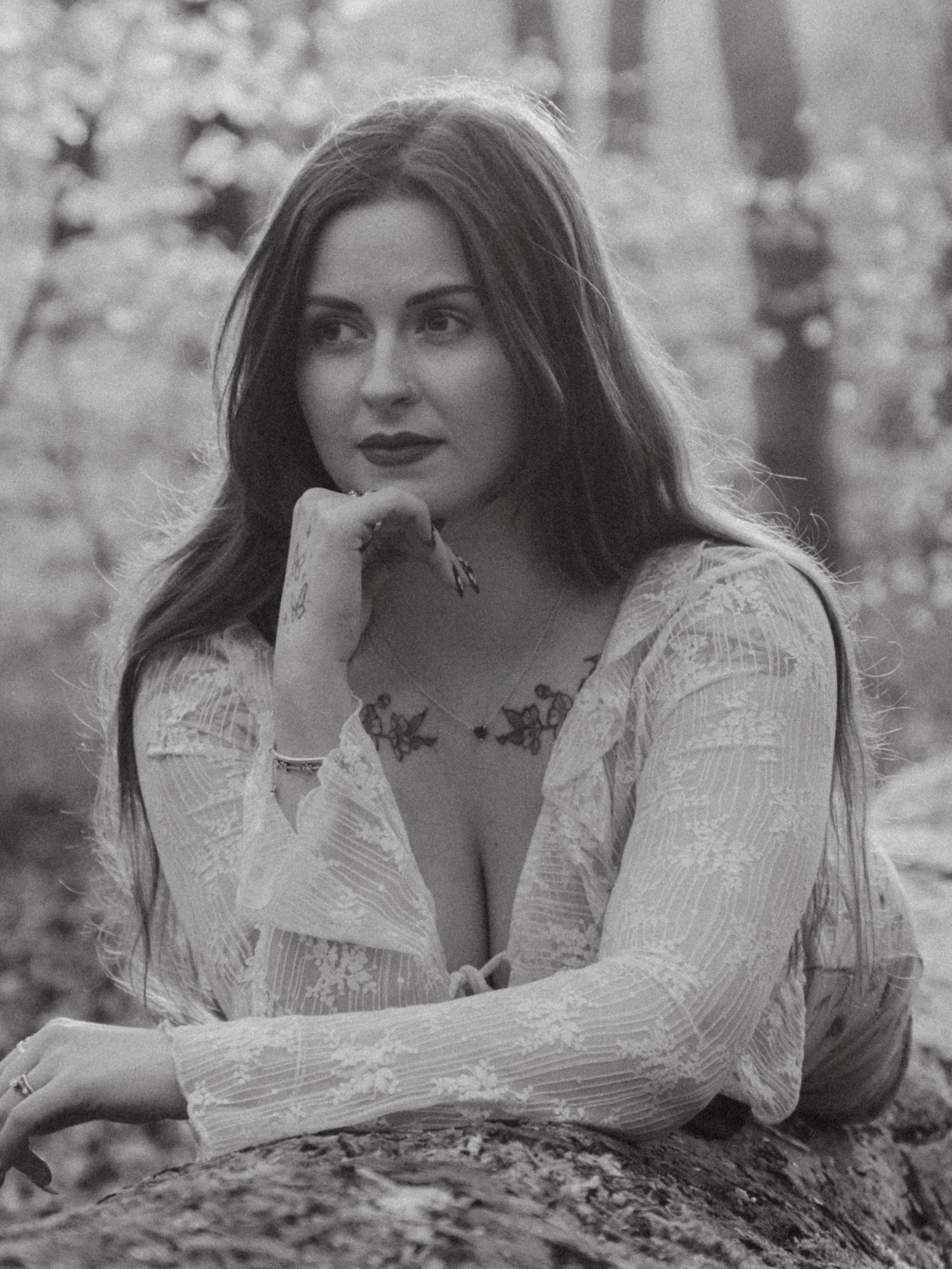 Black and white photo of a woman with long hair, wearing a lace dress, sitting outdoors on a tree branch. She is resting her chin on her hand and looking to the side.