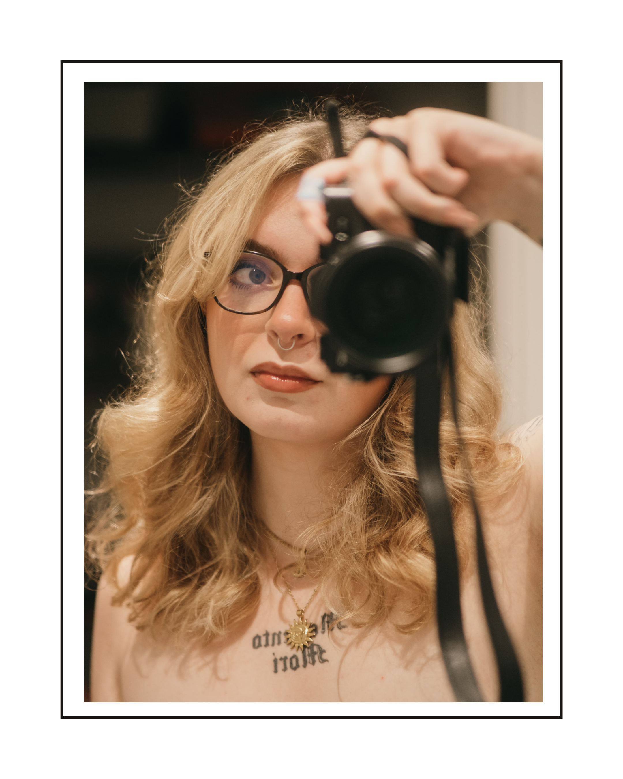 A young woman with long curly blonde hair, wearing black glasses, a septum piercing, and a gold sun necklace, taking a mirror selfie with a camera. She is wearing a light pink shirt with text partially visible.