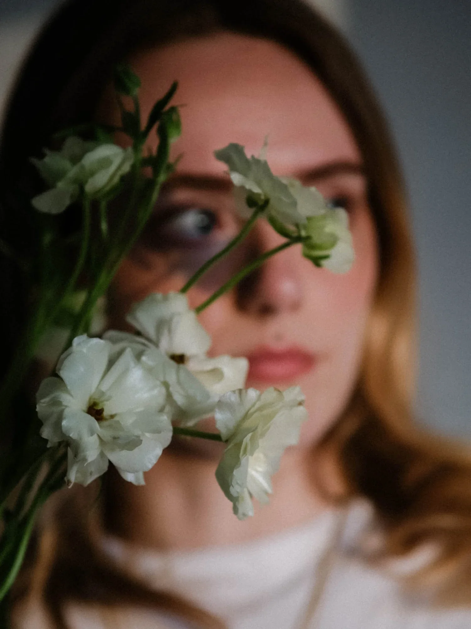 A woman with brown hair holding a bouquet of white flowers near her face, partially covering it, with a neutral gray background.