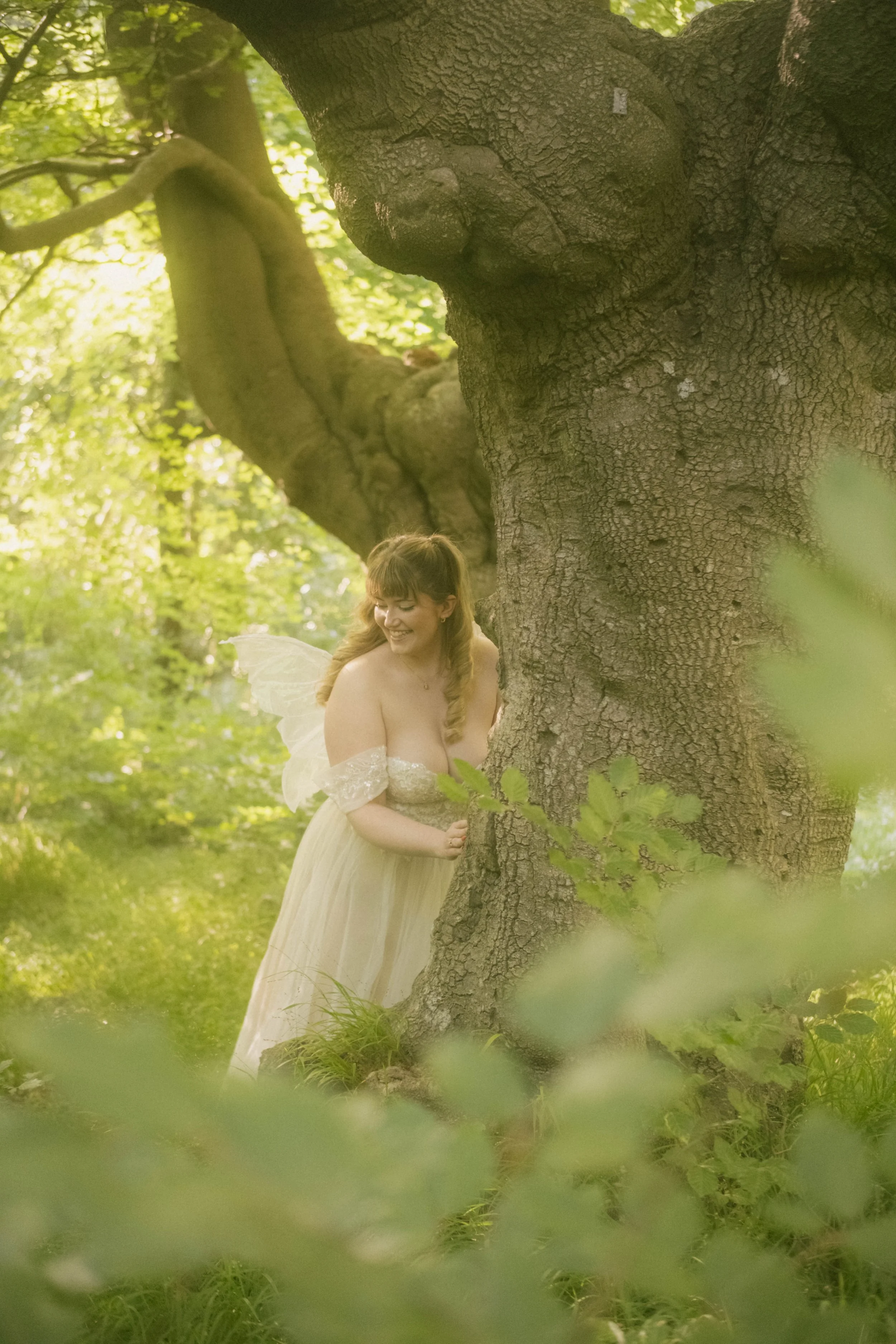 A woman dressed as a fairy with wings, standing behind a large tree in a lush, green forest, smiling and looking down.
