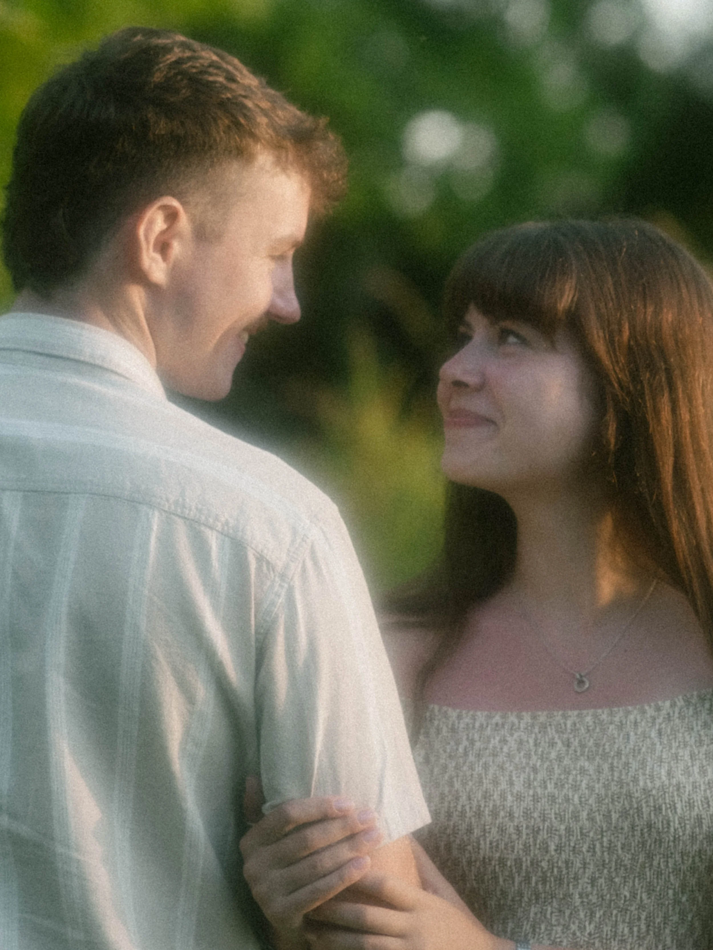 A young man and woman face each other outdoors, smiling gently, with blurred trees in the background.