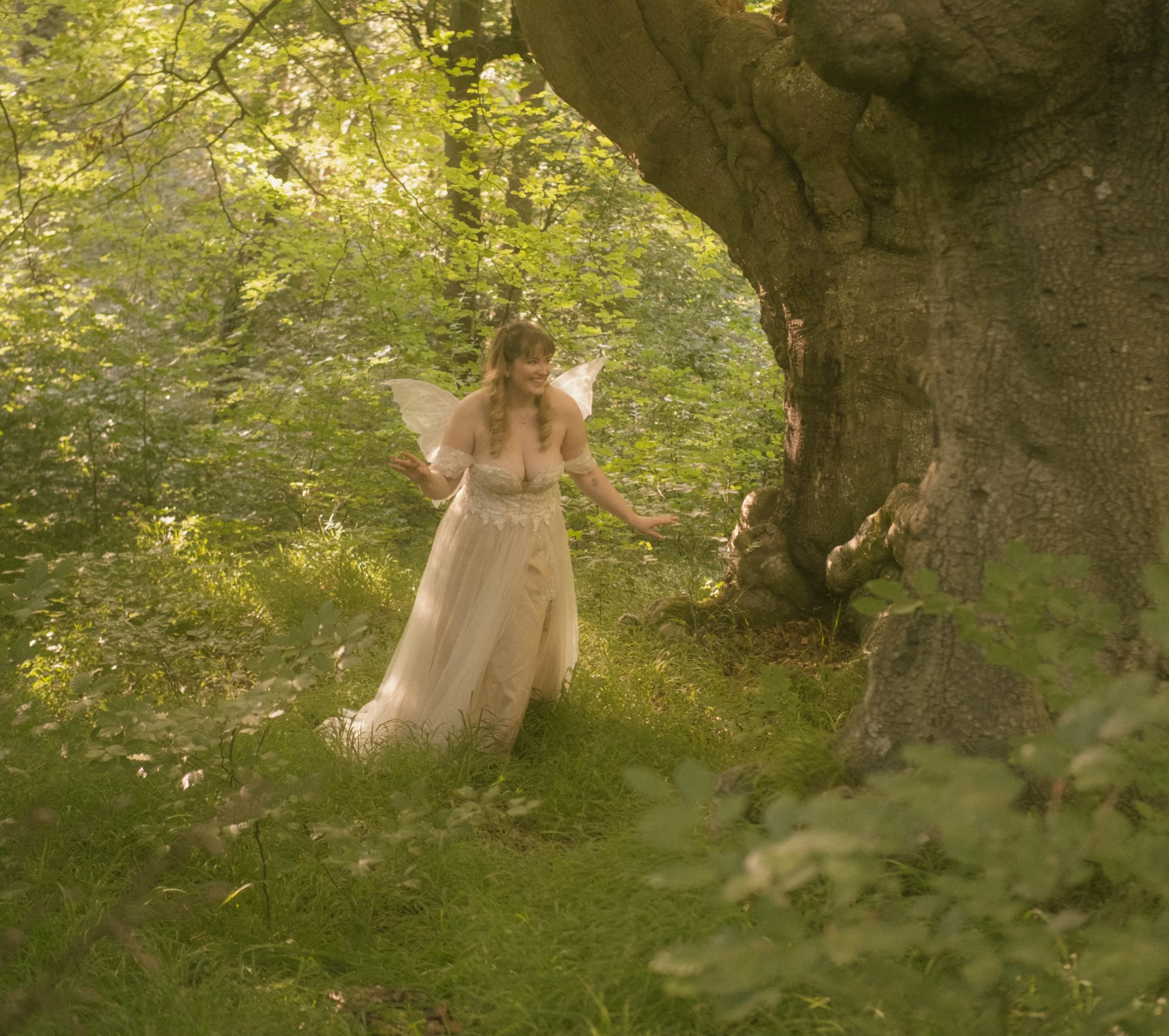A woman dressed as a fairy with butterfly wings standing in a lush green forest near a large tree trunk, sunlight filtering through the leaves.