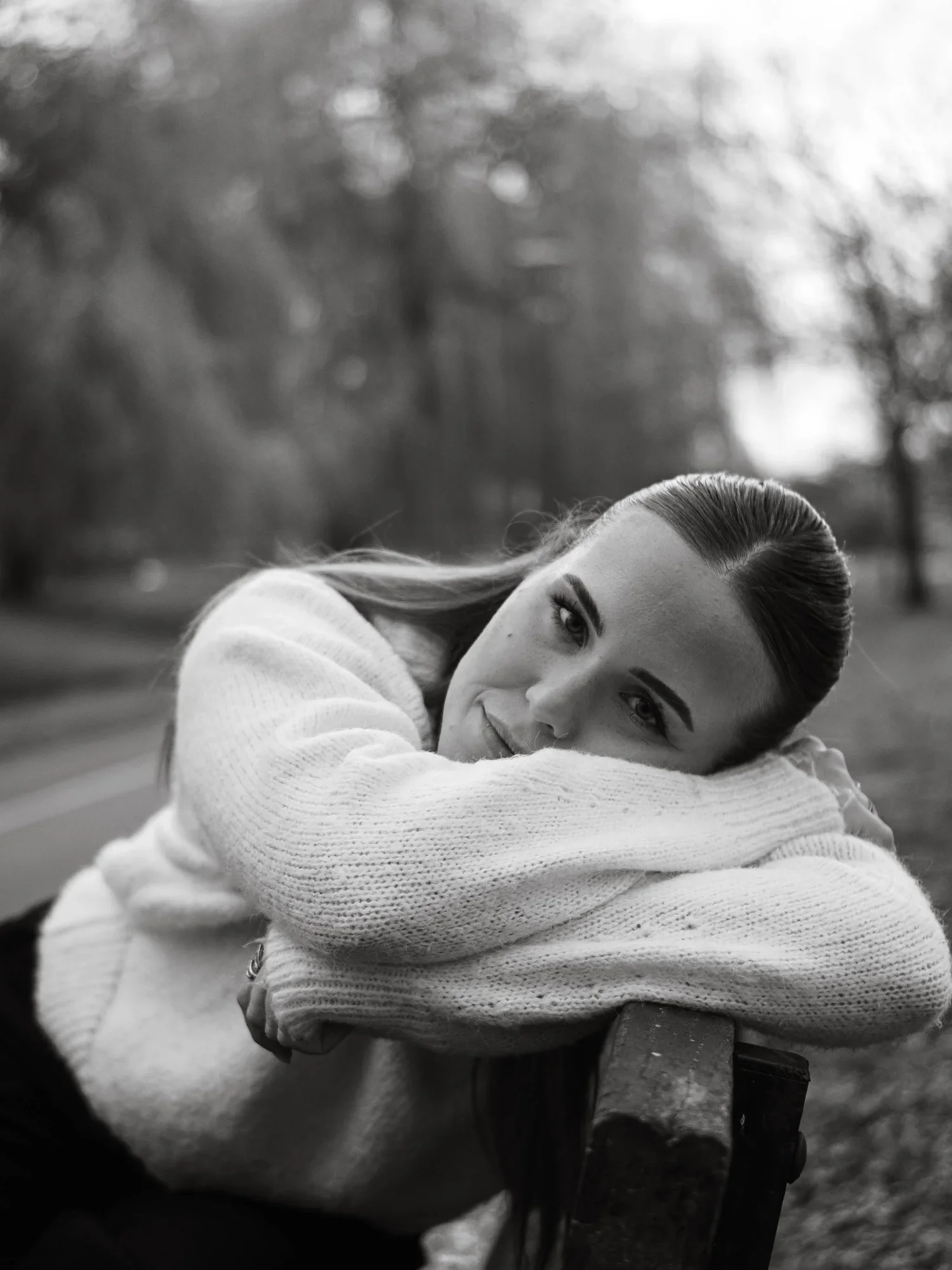 Black and white photo of a woman with long hair, resting her head on her arm on a table outdoors with blurred trees in the background.