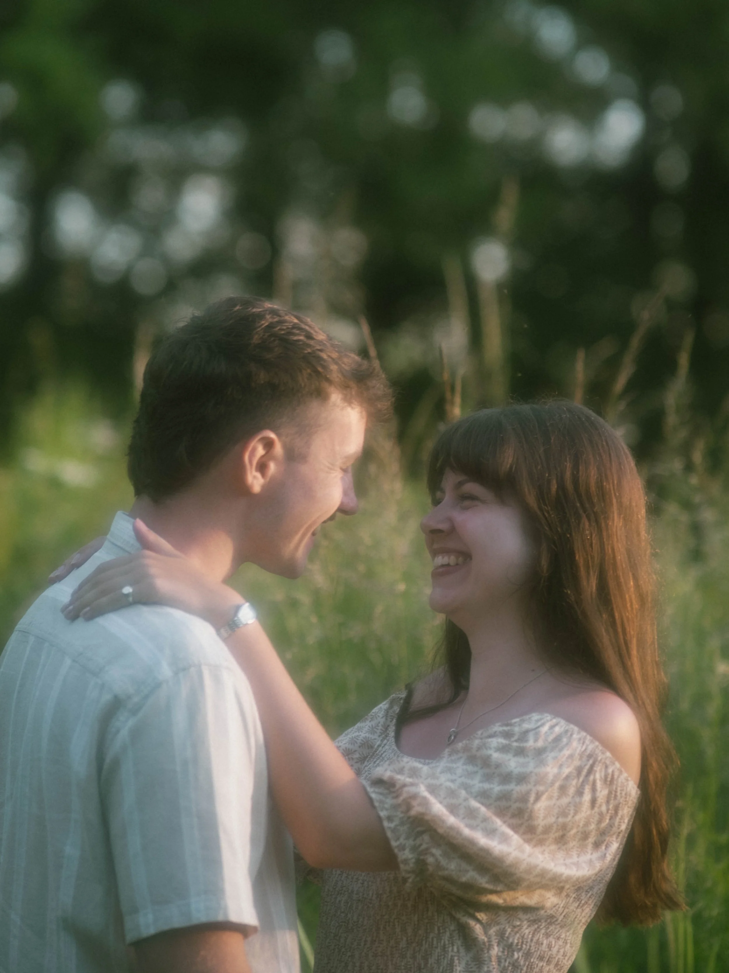 A man and a woman are facing each other, smiling and close together outdoors. The background shows trees and greenery.