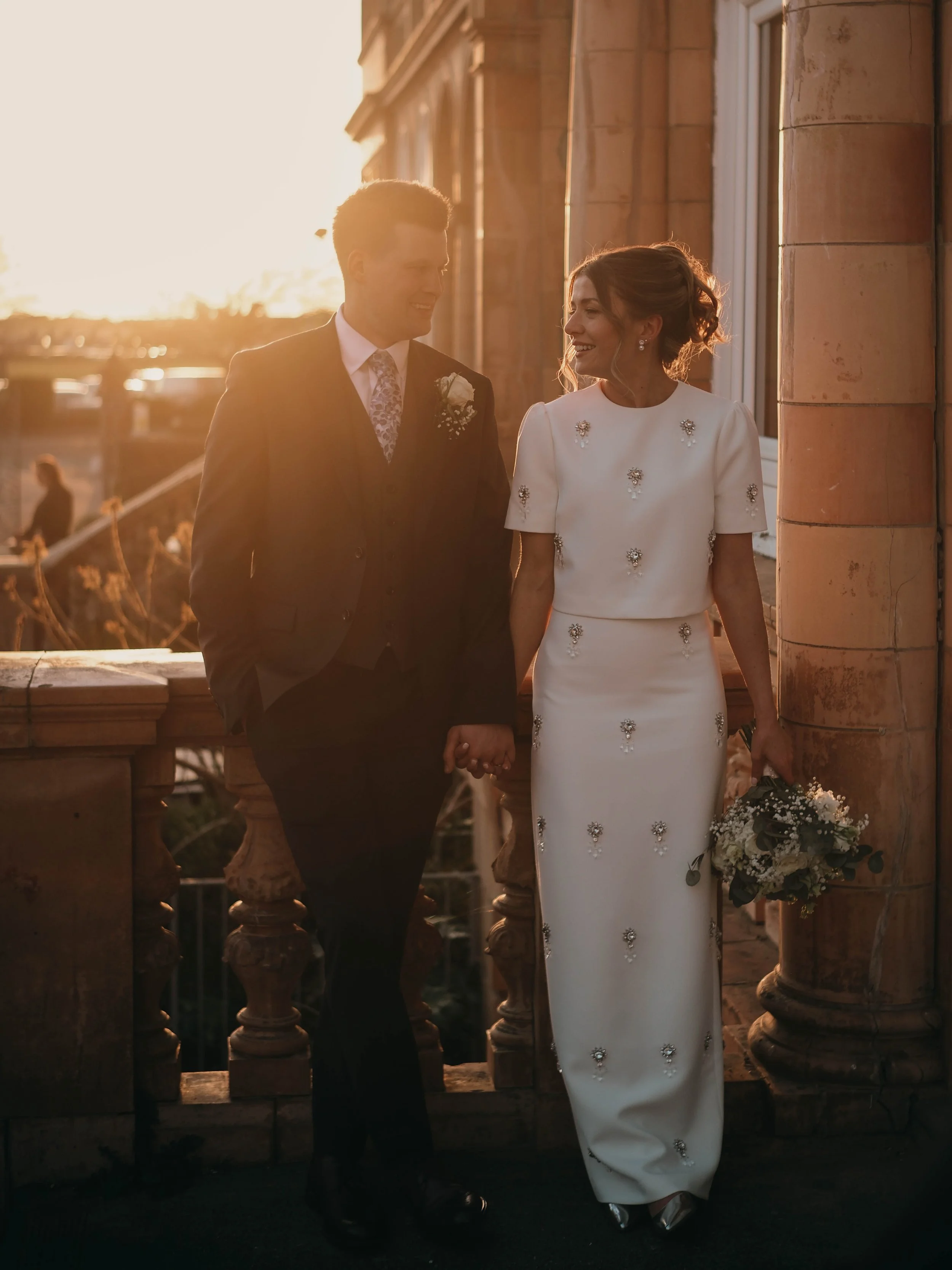A couple dressed in wedding attire standing outdoors at sunset, holding hands and smiling at each other. The man is wearing a suit with a floral tie and boutonniere, and the woman is in a white gown with floral embellishments, and holding a bouquet o