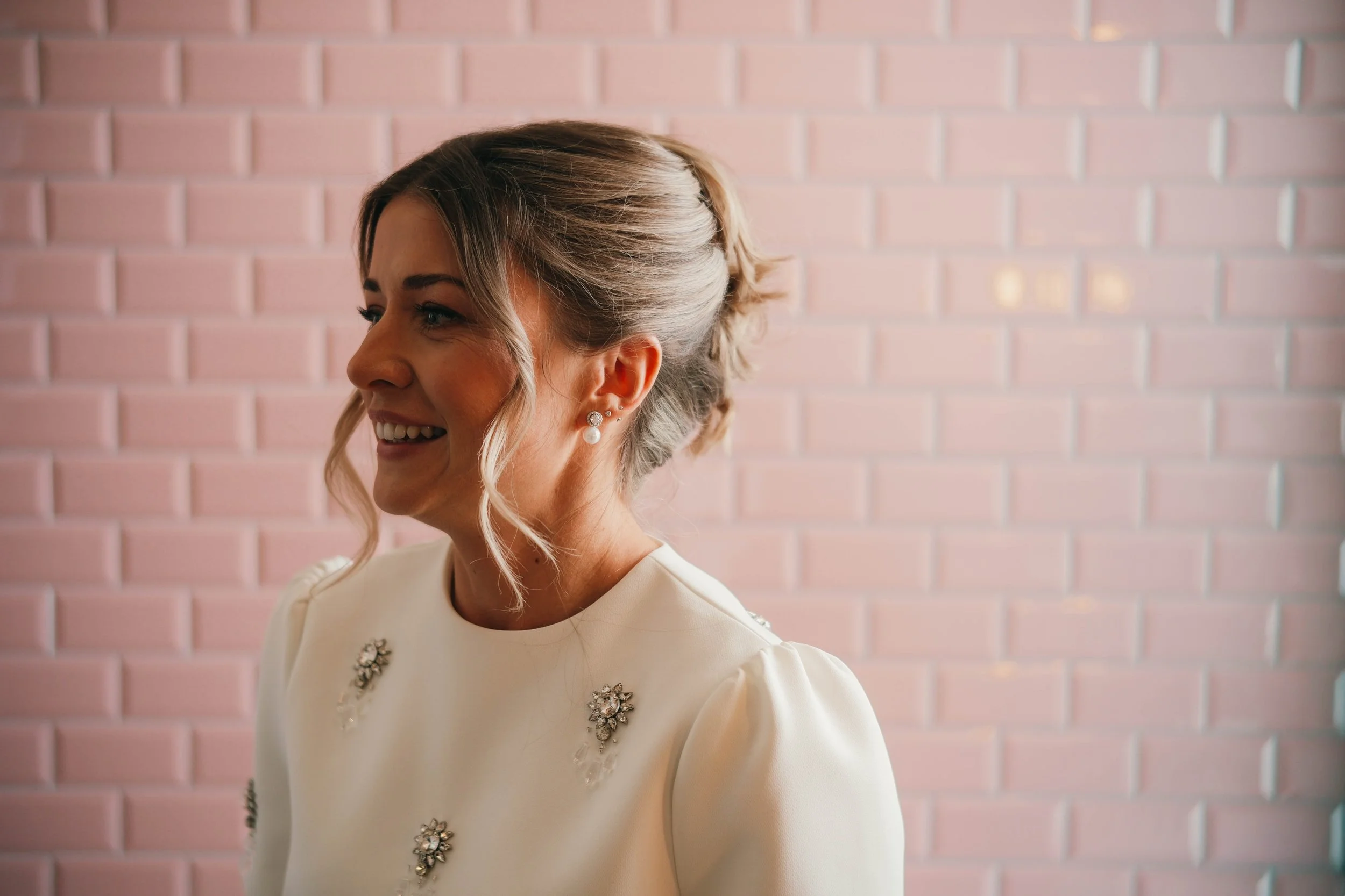 Side profile of a woman with styled gray hair, wearing pearl earrings, and a white top with floral embellishments, standing in front of a pink brick wall.