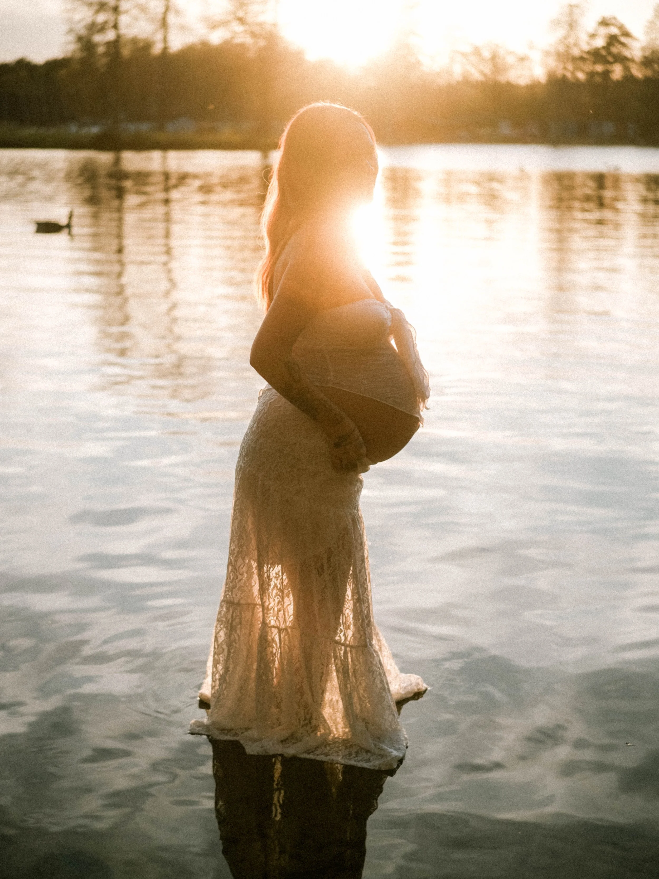A woman in a lace dress standing on a rock in a body of water during sunset, with trees and a bright sun in the background.