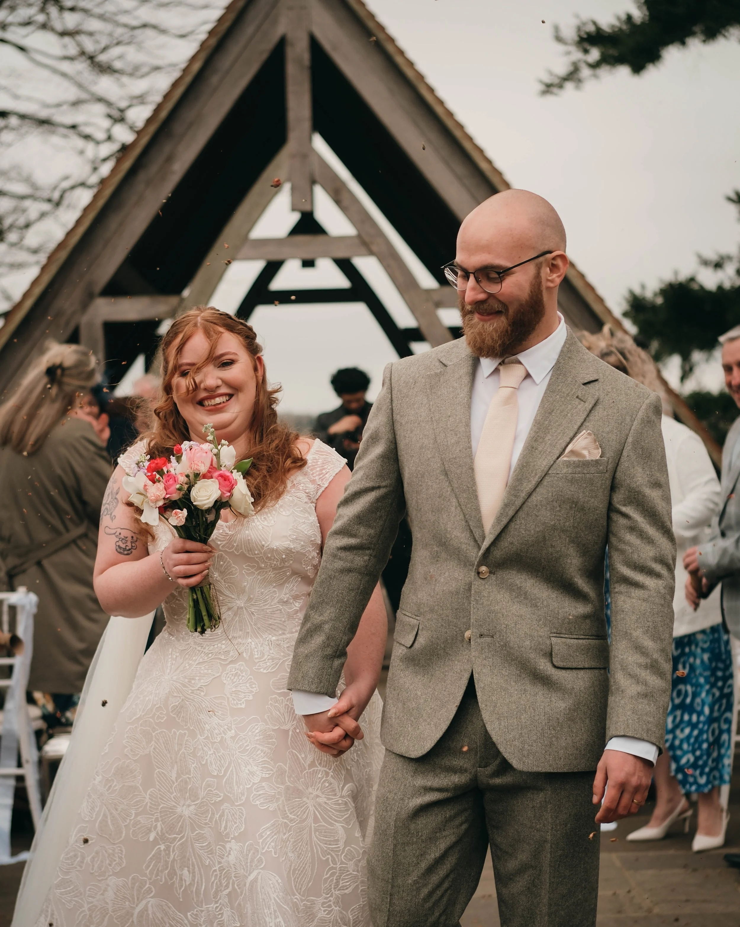 A newlywed couple holding hands during their outdoor wedding ceremony. The bride is holding a bouquet and wearing a lace wedding dress, while the groom is dressed in a gray suit with a light-colored tie. They are smiling and surrounded by friends and