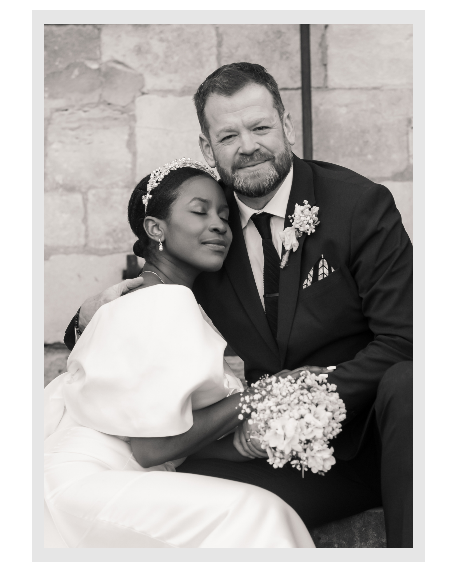 Black and white photo of a bride and groom, with the bride resting her head on the groom's shoulder and eyes closed. The bride is wearing a wedding dress and pearl earrings, and has a pearl headband. The groom is wearing a suit with a boutonniere and a pocket square. They are sitting close to each other, holding a bouquet of flowers. The background features a brick wall.