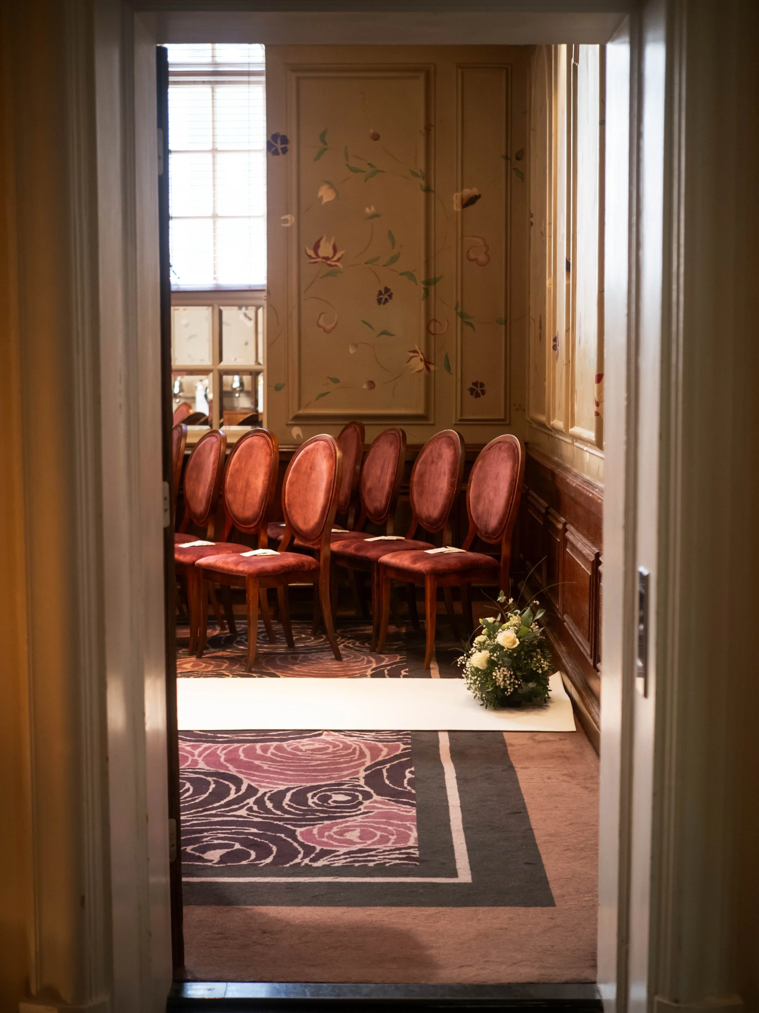 View through a doorway into a room with pink velvet chairs arranged in a row, a floral wall design, a window letting in natural light, and a floral arrangement on the floor.