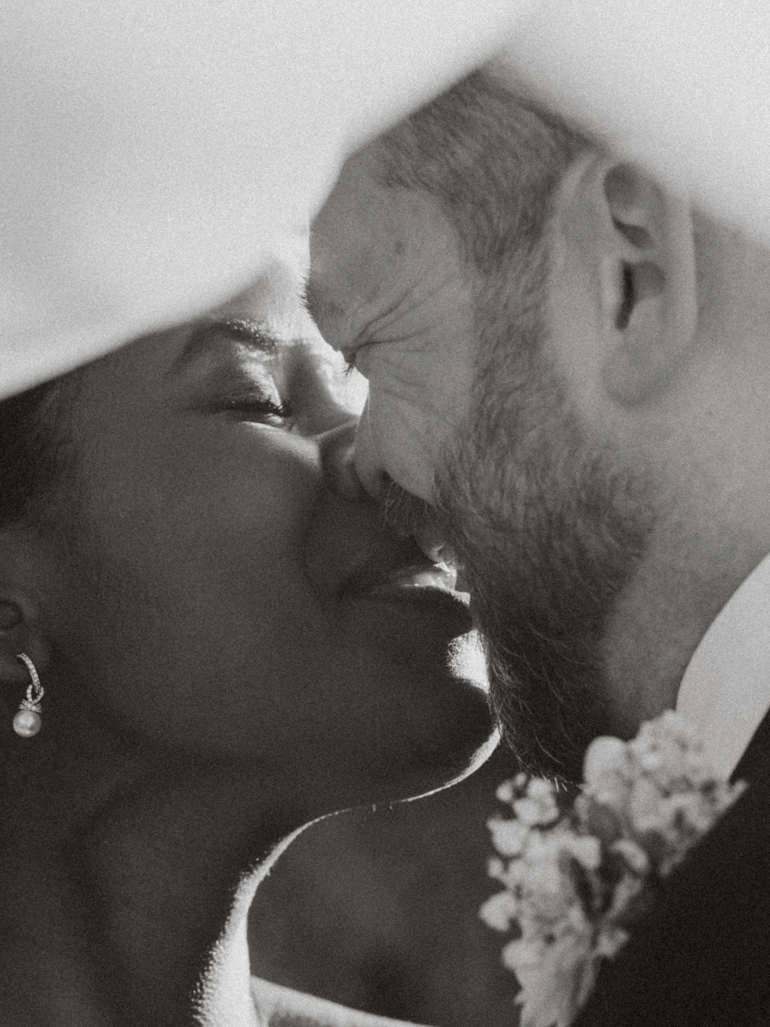 A black and white close-up photograph of a man and woman sharing a kiss, with flower petals and blurred background.