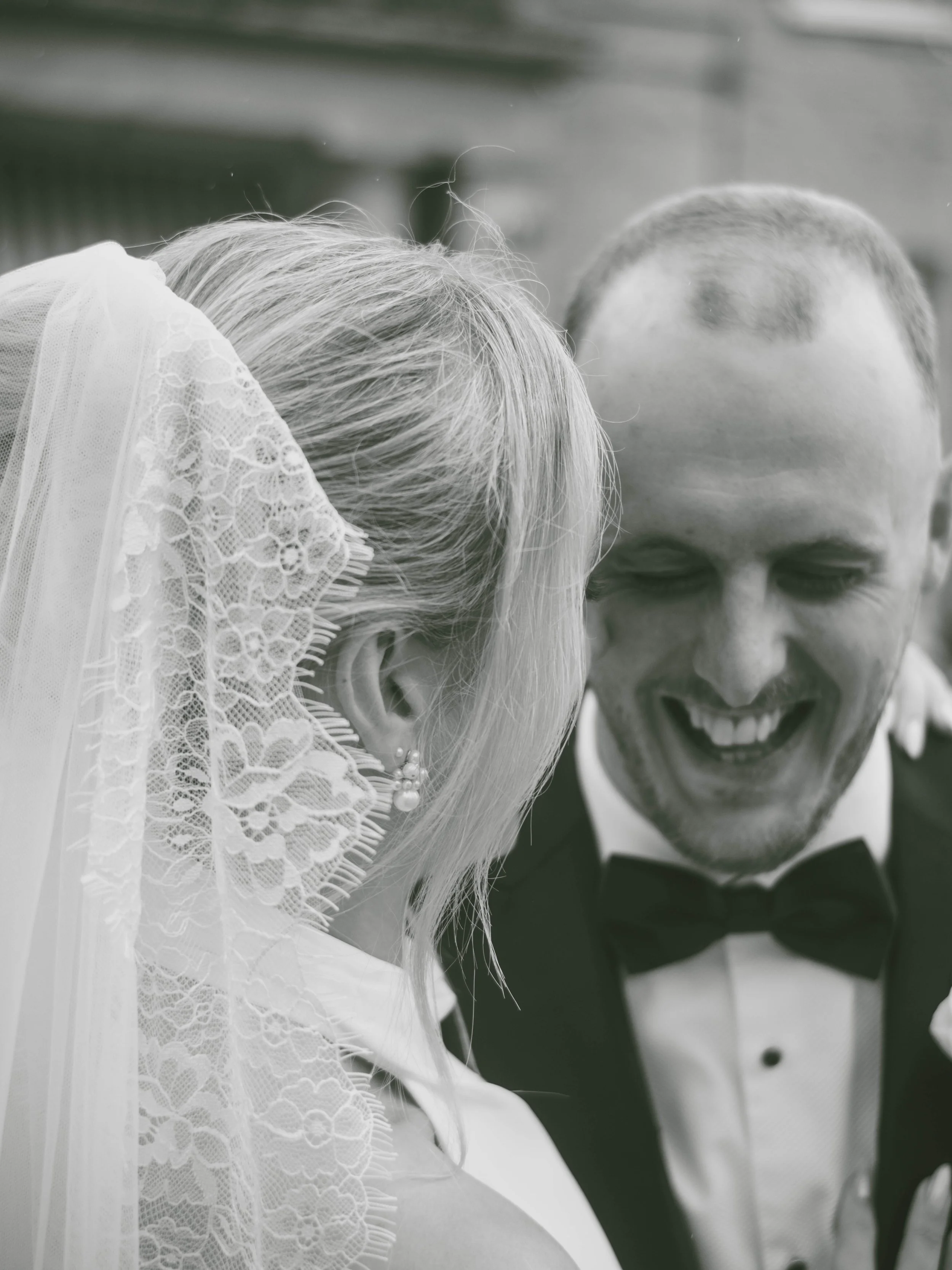 A black-and-white photo of a bride and groom. The bride's head is close to the groom's face, and the groom is smiling with his eyes closed. The bride is wearing a lace veil and a pearl earring.