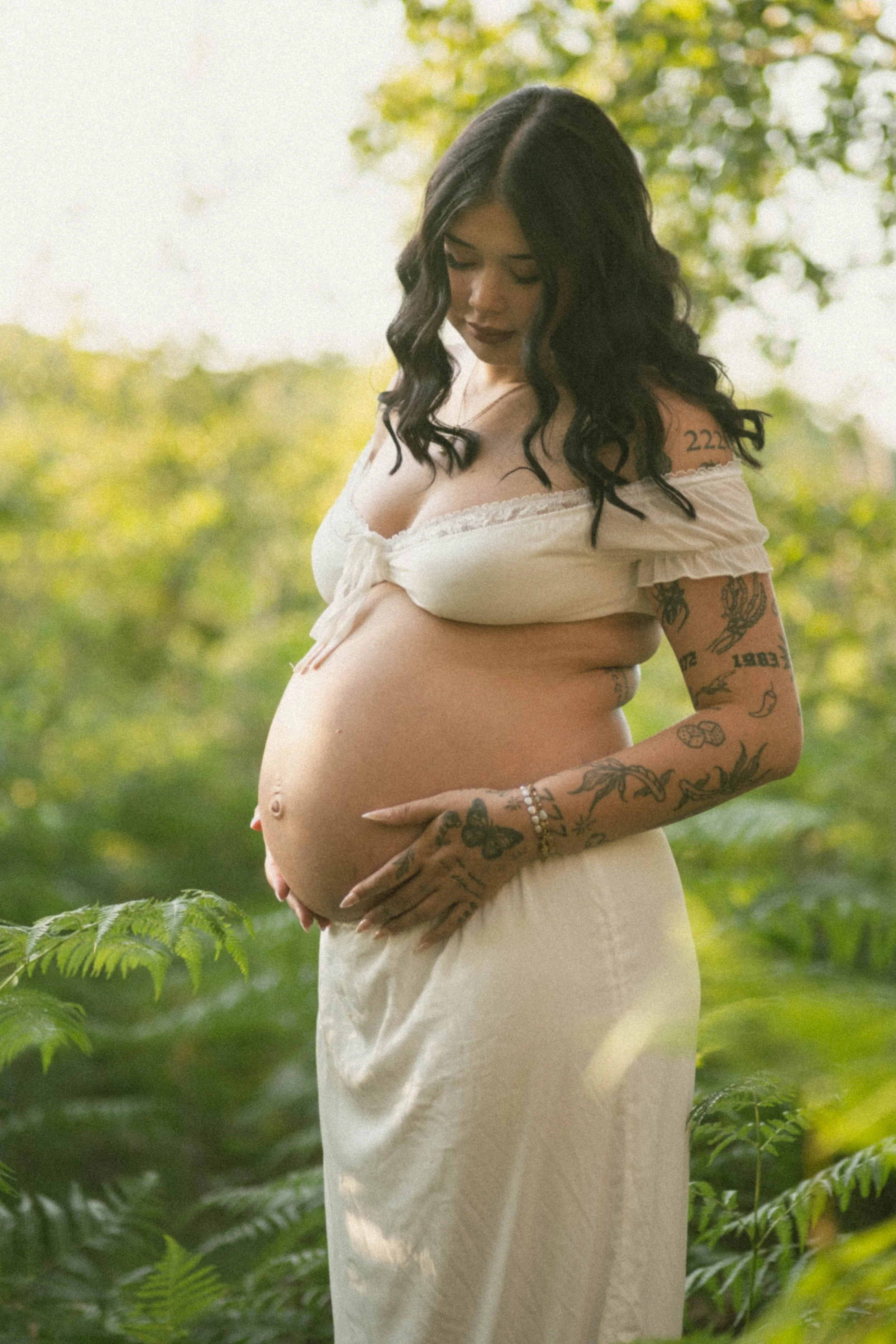A pregnant woman with dark wavy hair, tattoos on her arms, wearing a cream off-shoulder dress, standing outdoors among green plants and trees, gently cradling her belly.
