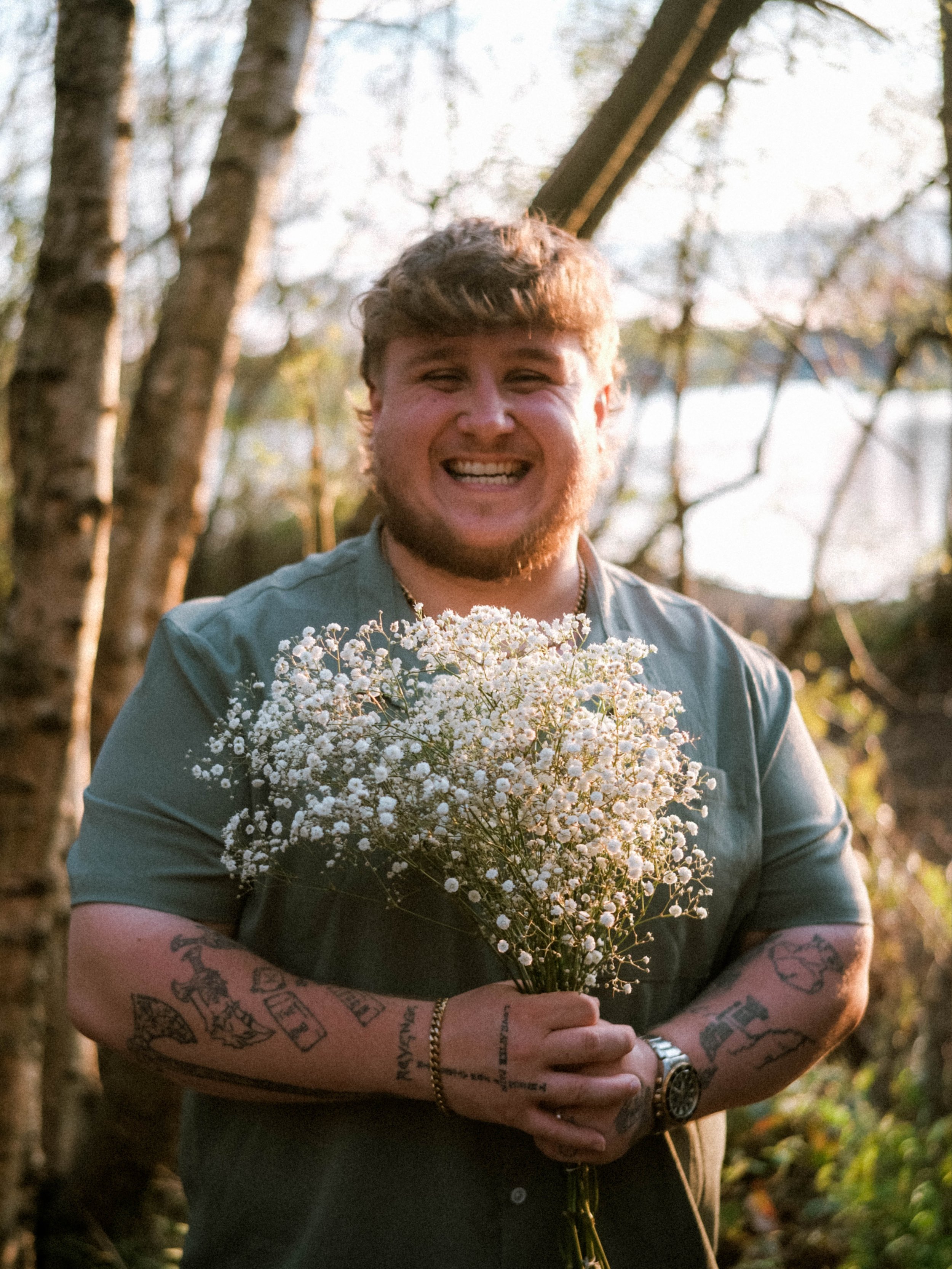 A smiling young man with tattoos on his arms holding a bouquet of white flowers outdoors, surrounded by trees and sunlight.