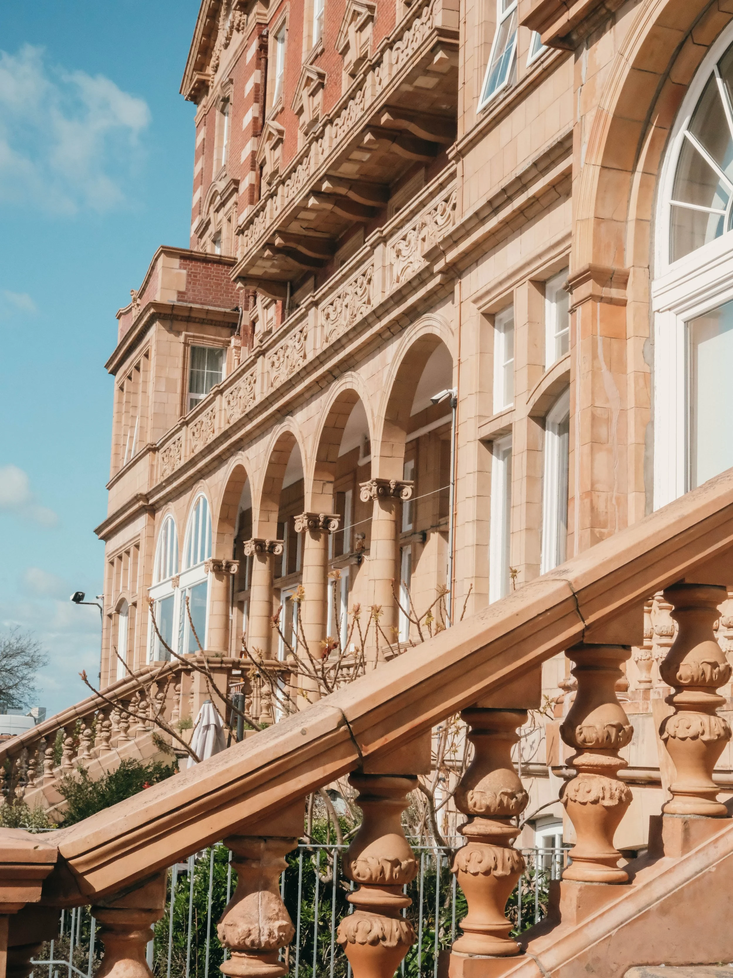 A close-up view of a historic building with ornate architectural details, arched windows, and a balustrade in the foreground, with a bright blue sky and some clouds in the background.