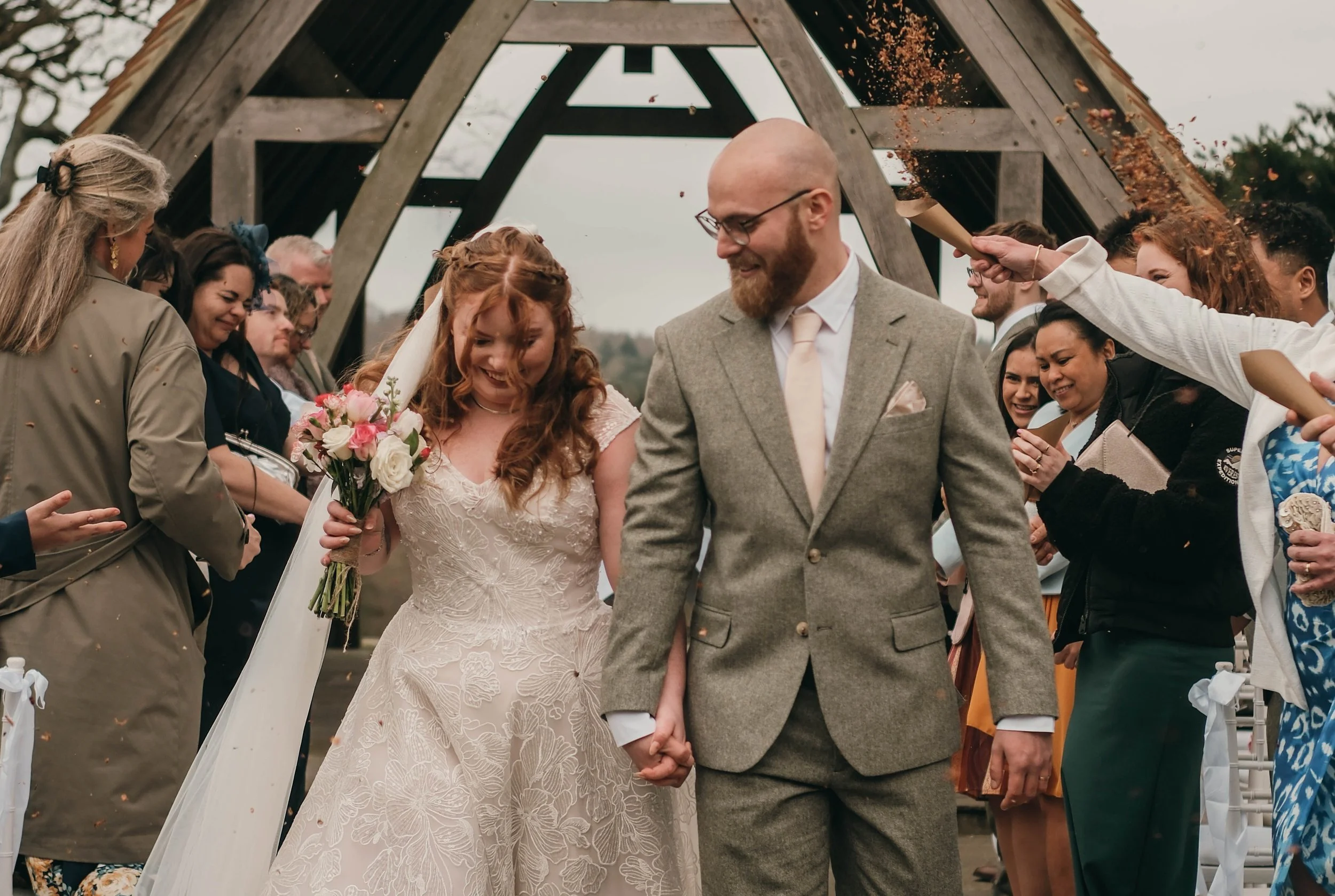 A wedding ceremony outside with a French bulldog dressed in a tuxedo sitting on a person's shoulders, surrounded by guests. The bride and groom are holding hands, smiling, and the bride is holding a bouquet of flowers.