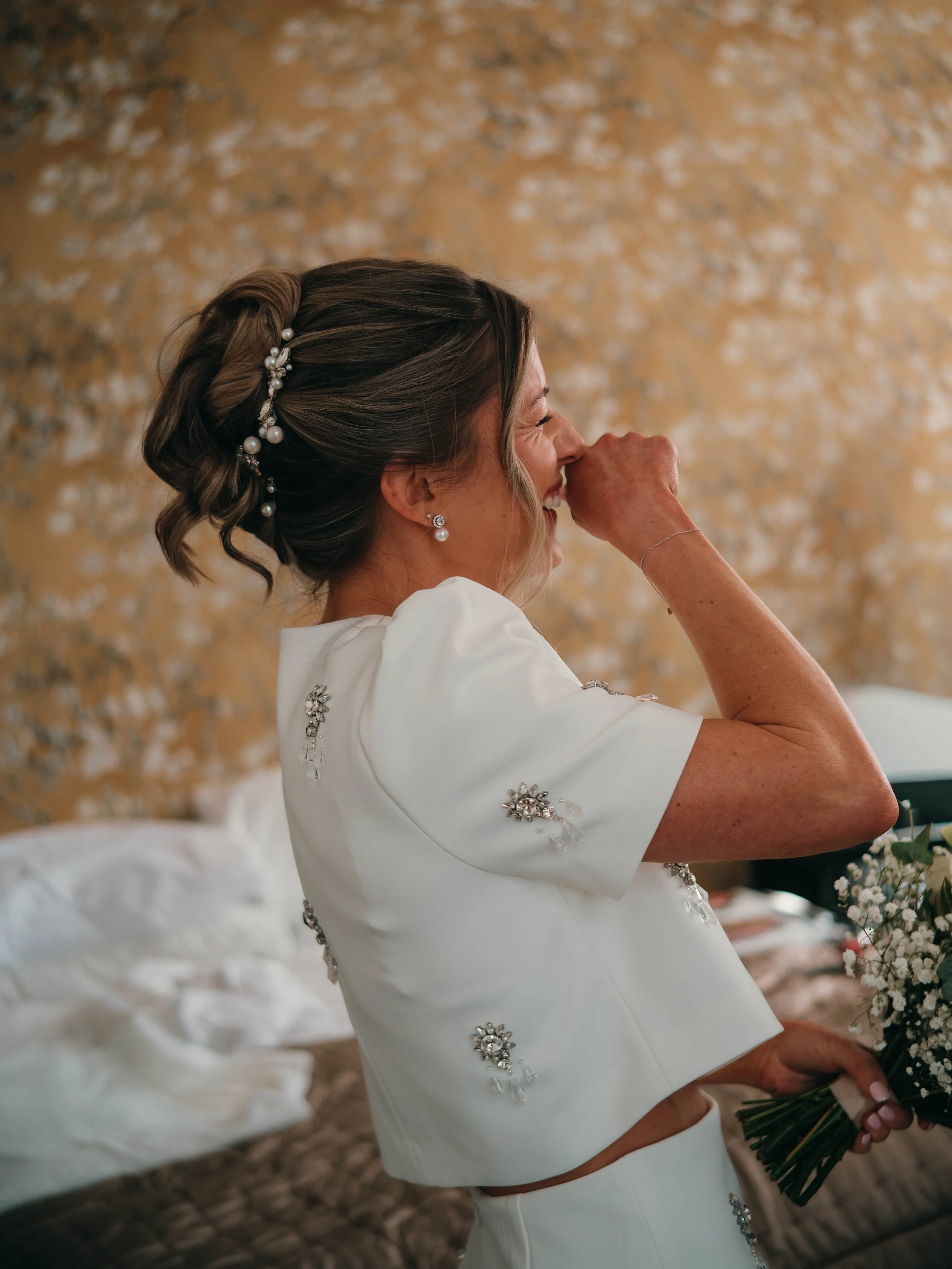 A woman with styled brown hair decorated with pearl hairpins, wearing earrings and a white dress with floral embellishments, holding a bouquet of flowers, smiling and appearing emotional indoors with a blurred background.