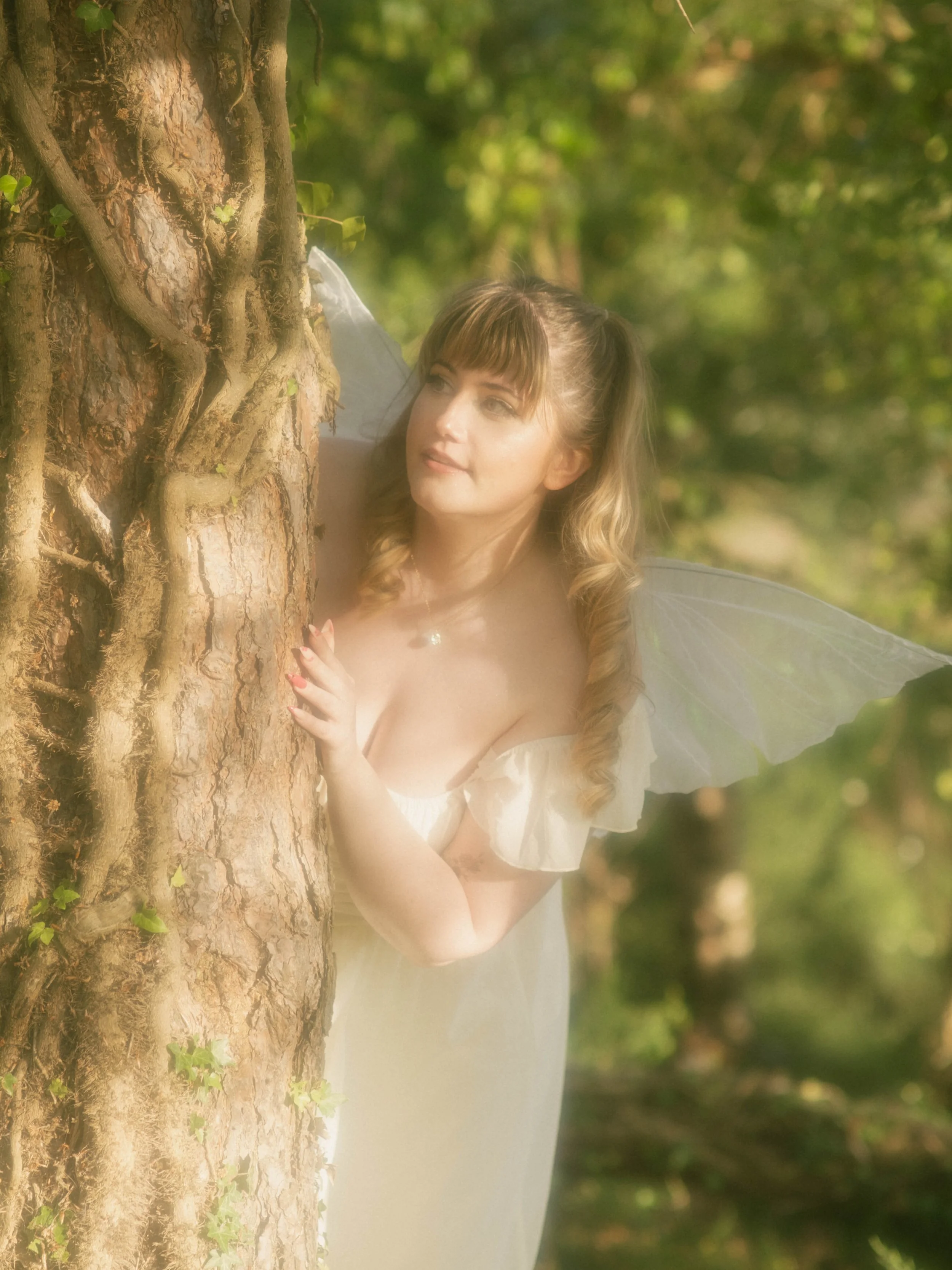 A fairy with blonde hair in pigtails, wearing a white dress and translucent wings, leaning against a large tree with textured bark and green ivy, in a lush forest with sunlight filtering through the leaves.