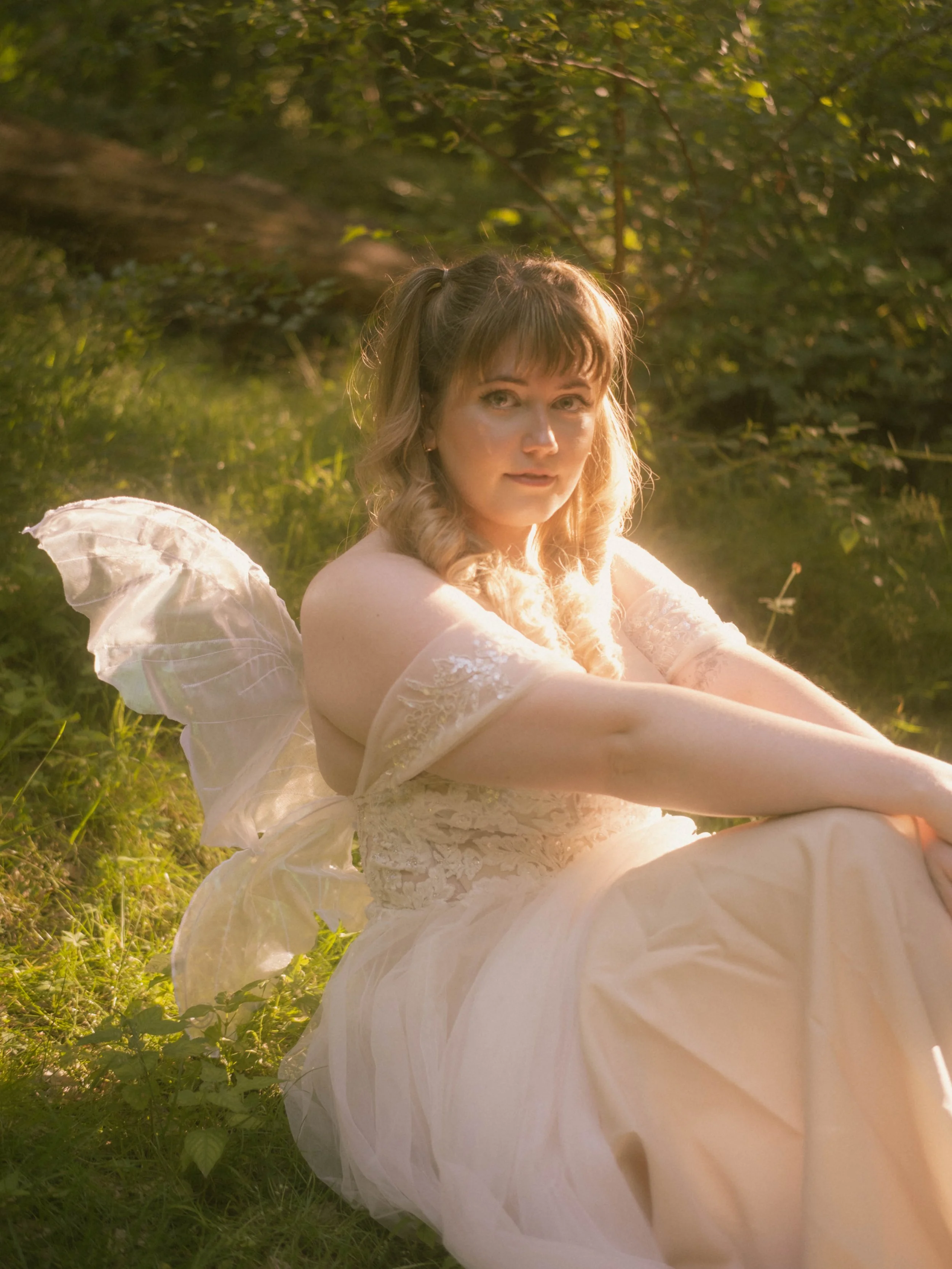 A woman with light brown hair, wearing a white lace dress with puffy sleeves, sitting outdoors on grass surrounded by green foliage, illuminated by warm sunlight.