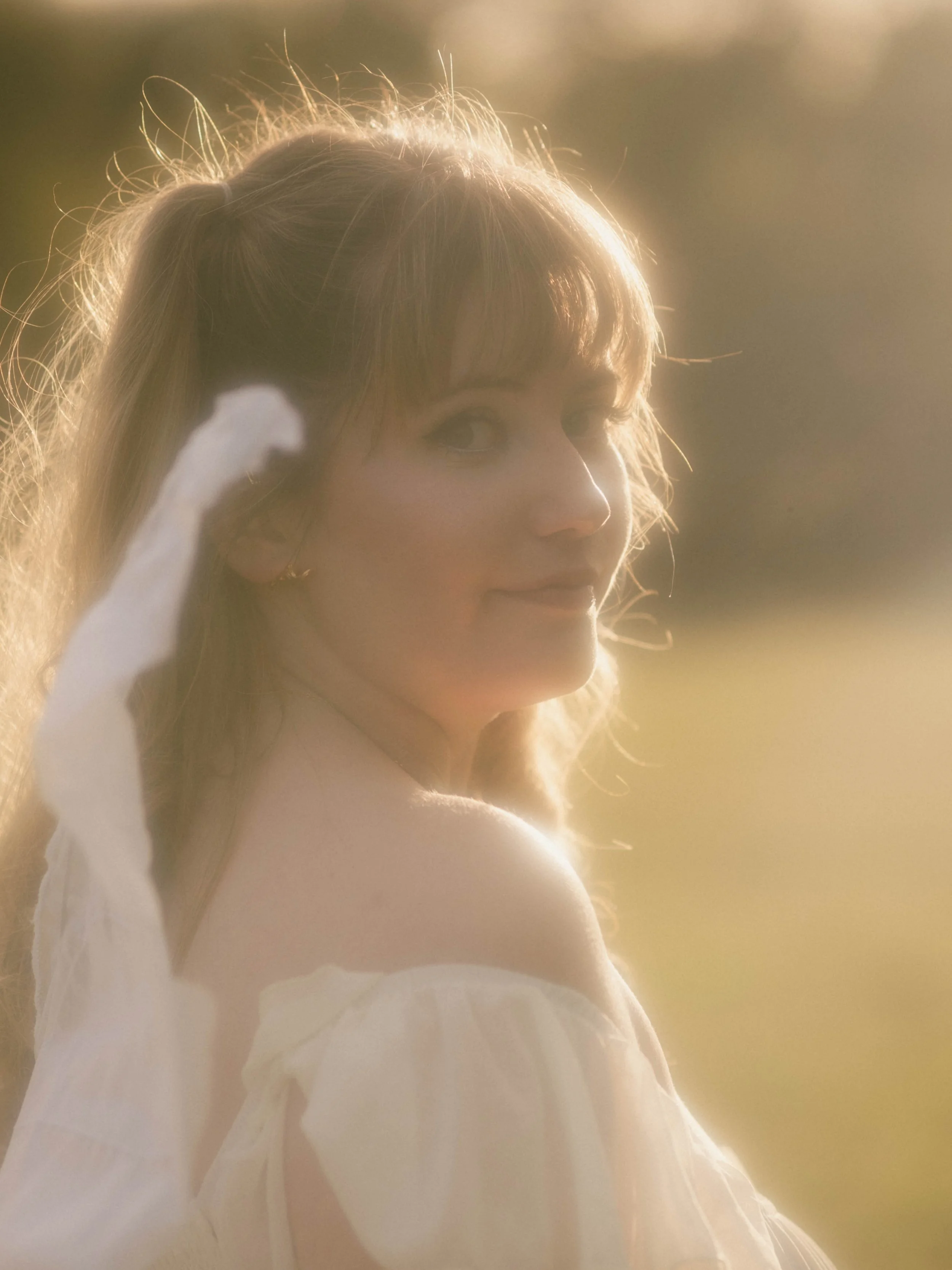 A woman with light brown hair and a gentle smile, partially covered in a white dress, looking back over her shoulder in warm sunlight.