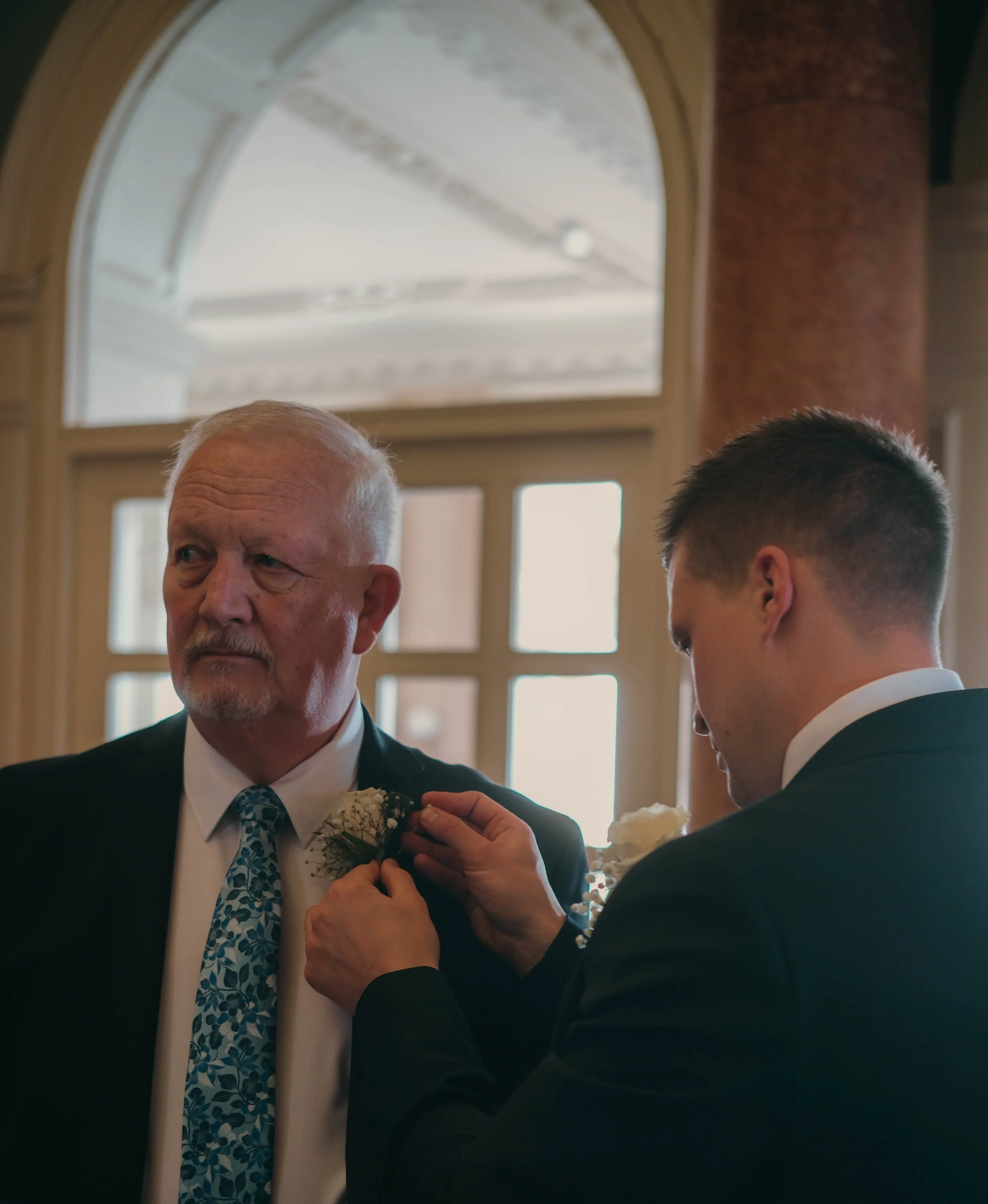 An older man and a younger man are dressing the older man in a boutonnière during a wedding or formal event inside a building with a large arched window in the background.