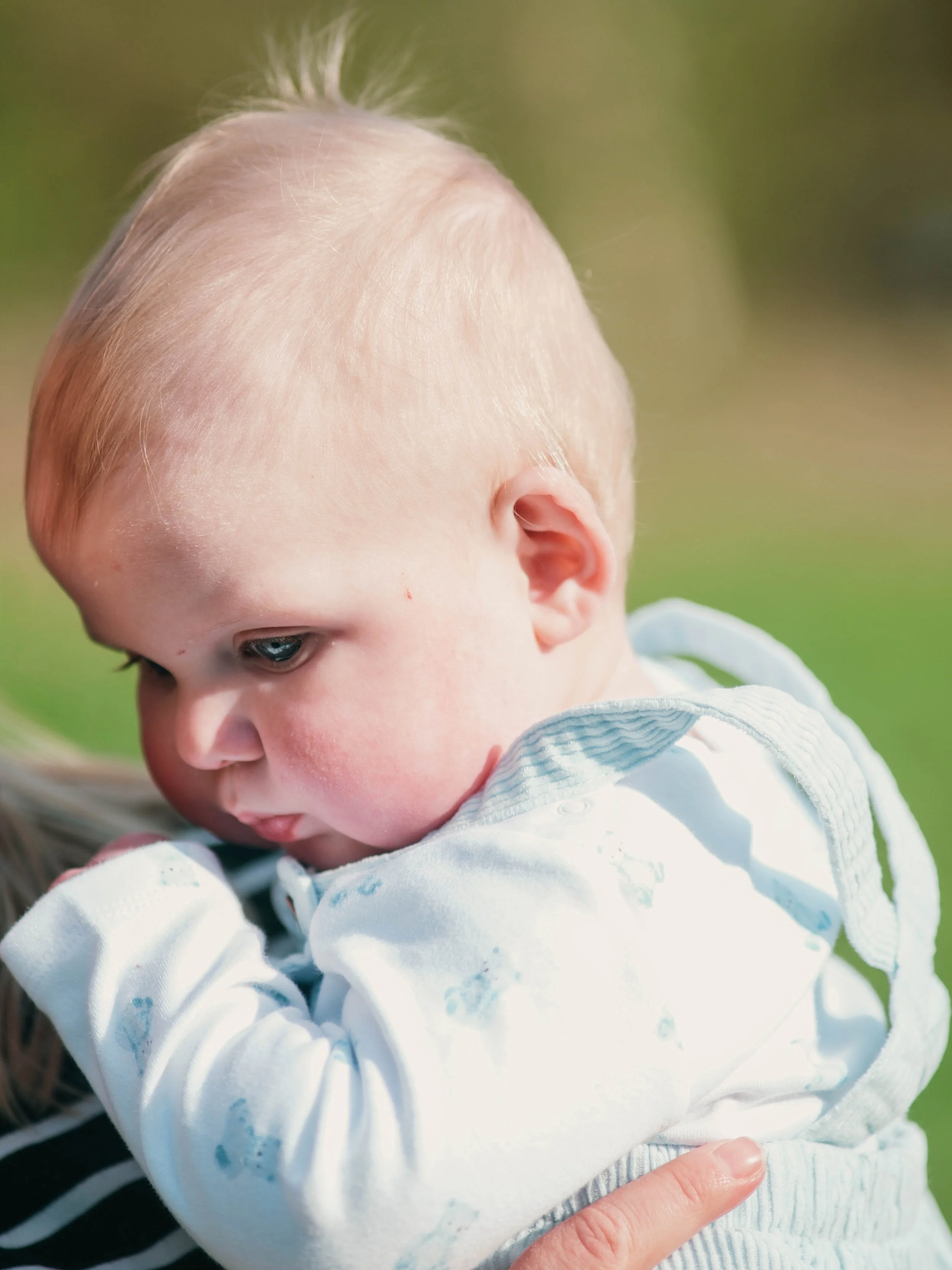 Close-up of a young child's head and face with blonde hair, wearing a white jacket, outdoors on a sunny day.