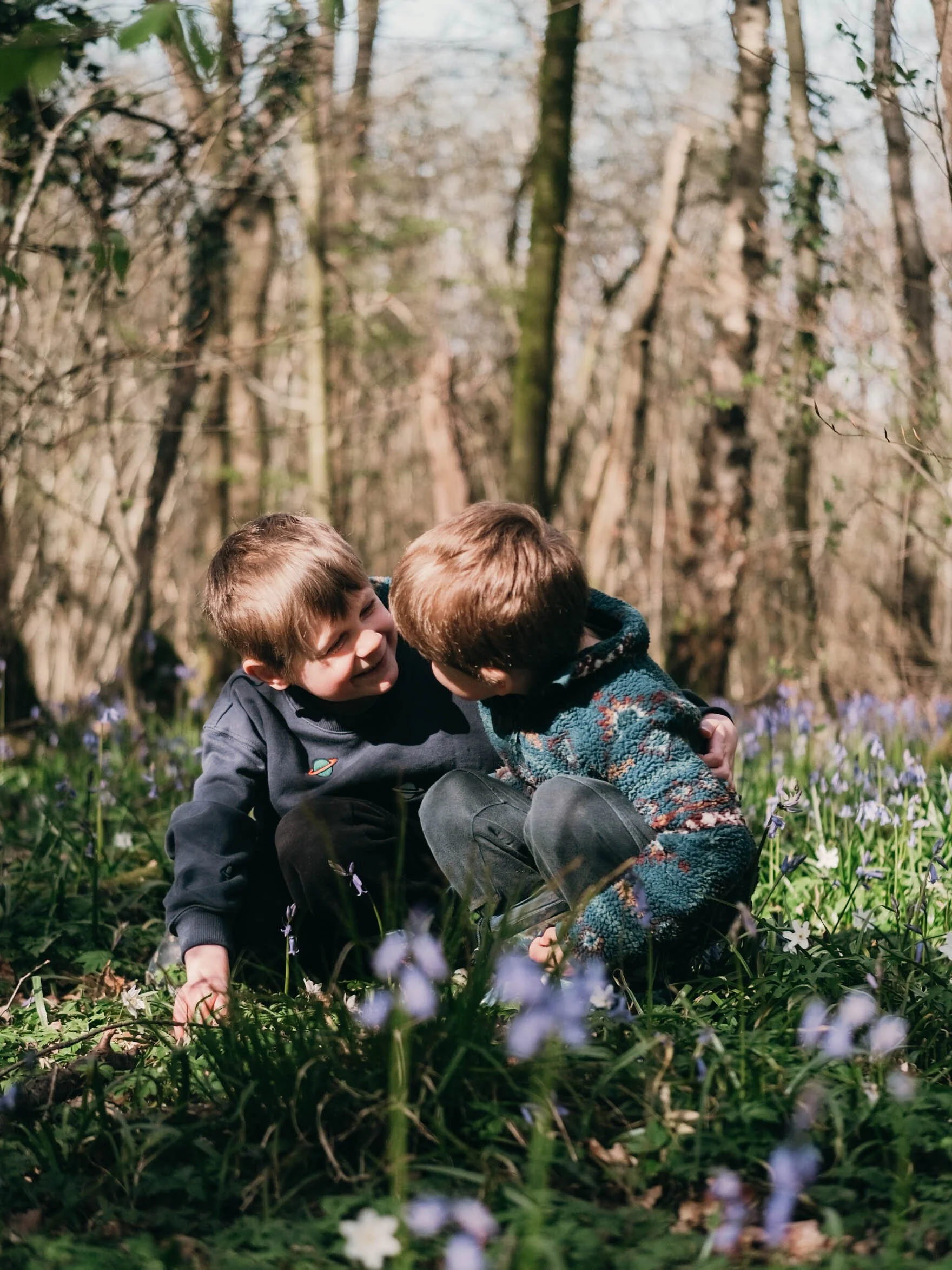 Two children are playing and hugging in a wooded area with blooming flowers on the ground.