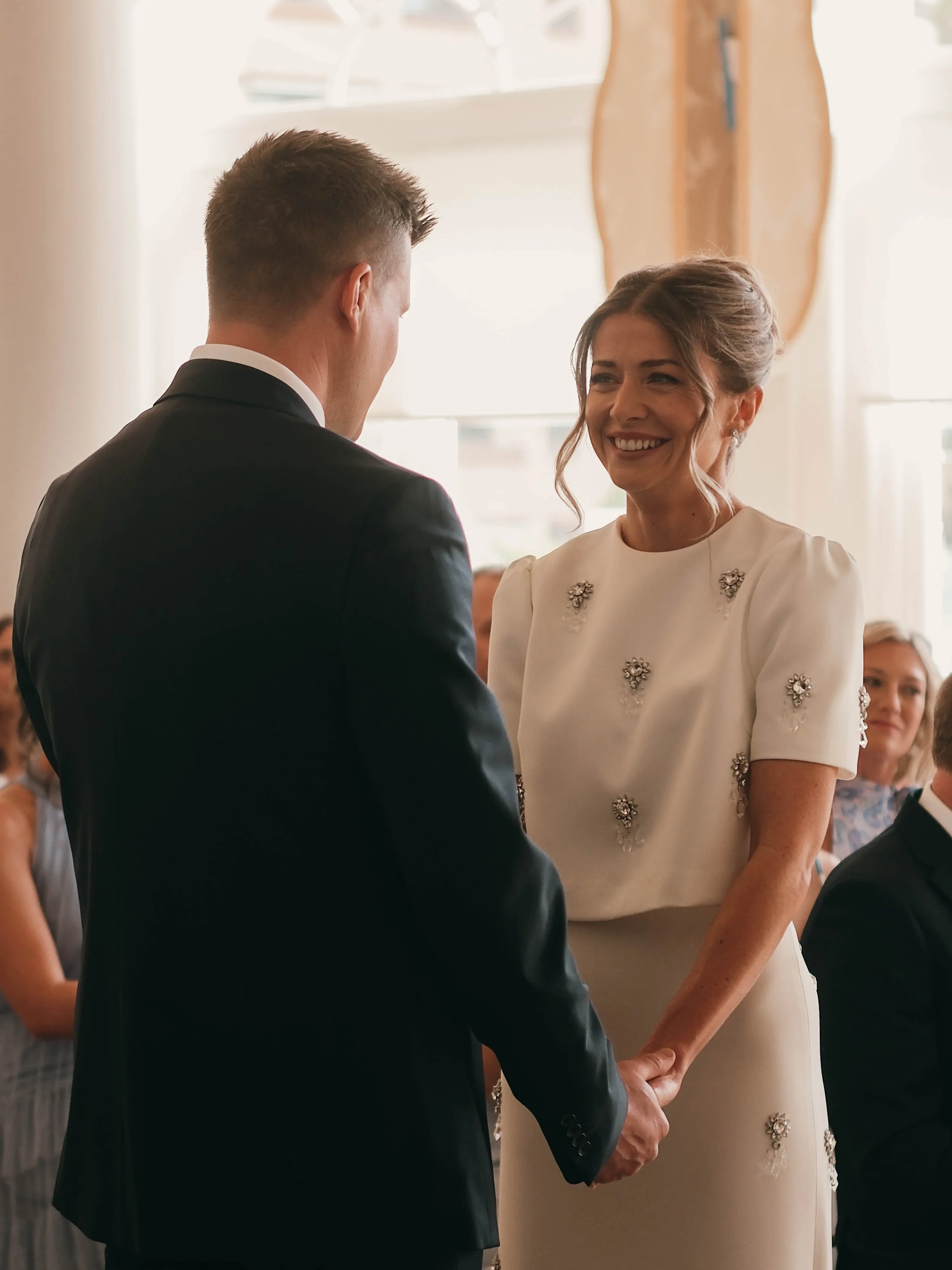 A groom and bride hold hands during their wedding ceremony, smiling at each other, with guests seated in the background.