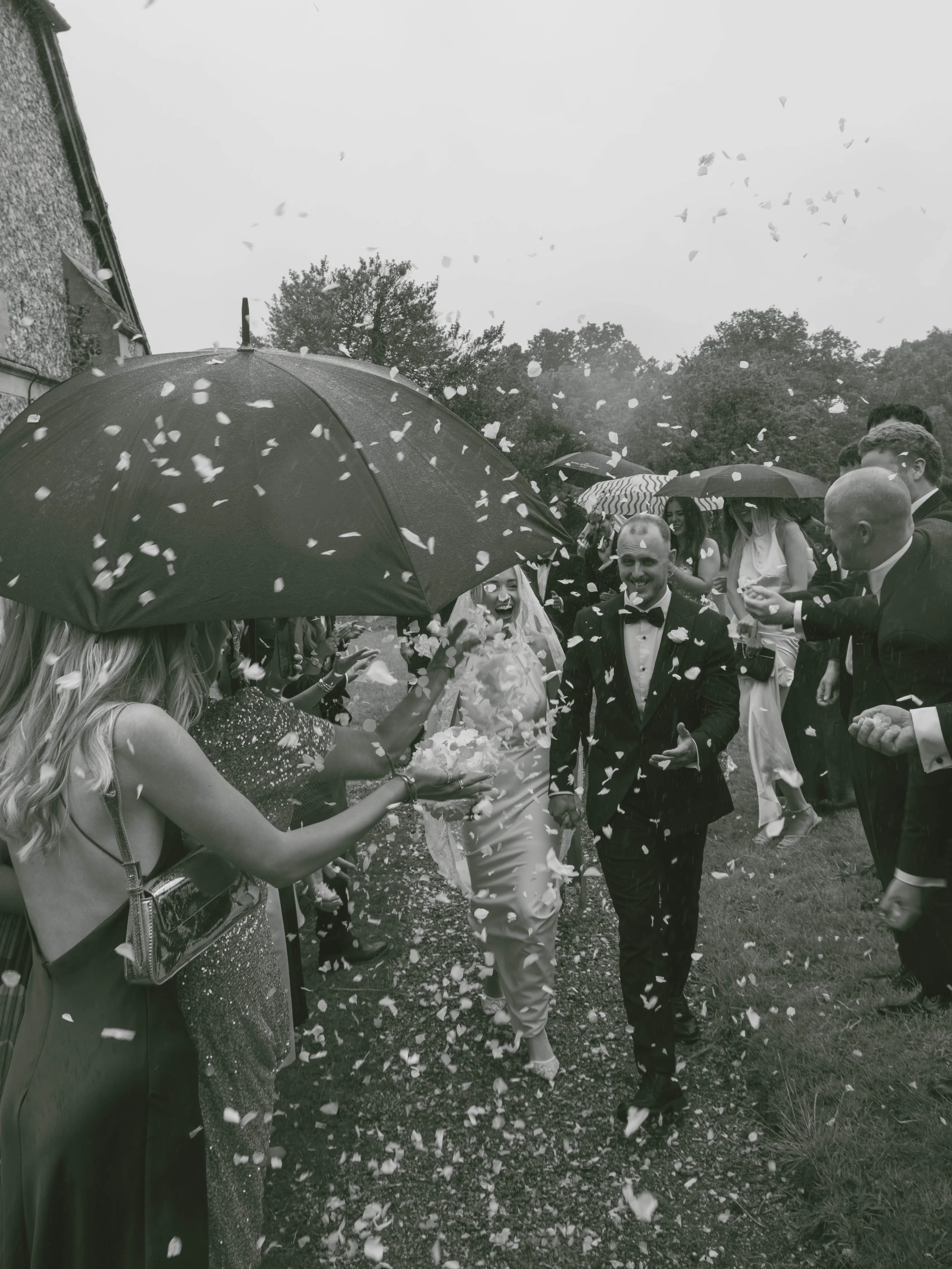 A wedding celebration outdoors with guests throwing flower petals, some holding umbrellas, on a rainy day.