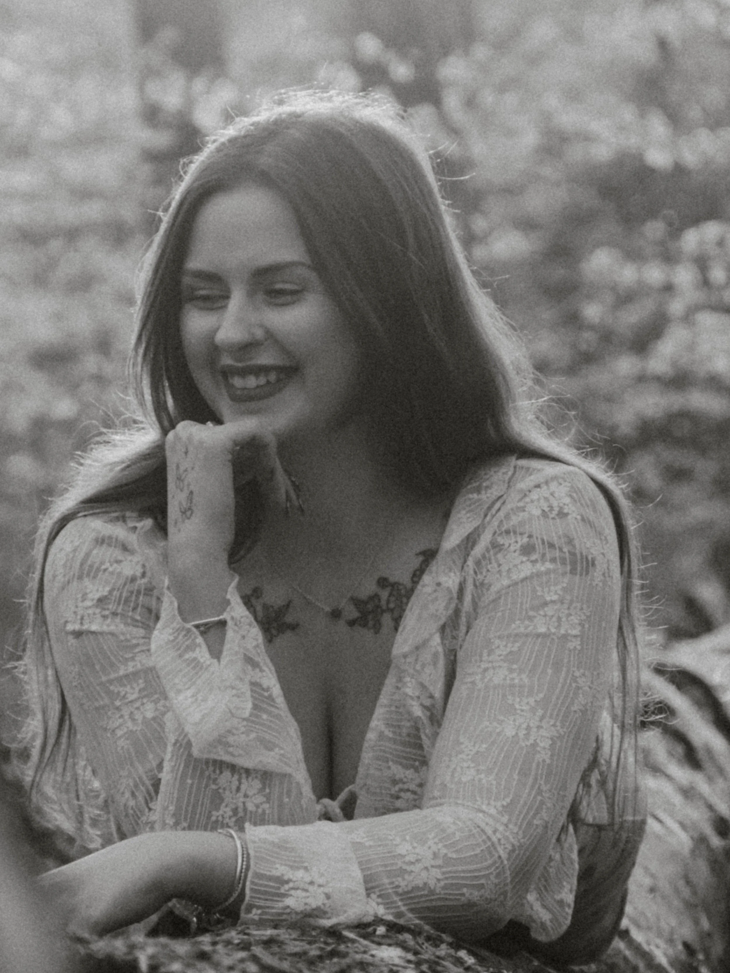 A woman with long hair smiling and resting her chin on her hand, outdoors with a blurred background.