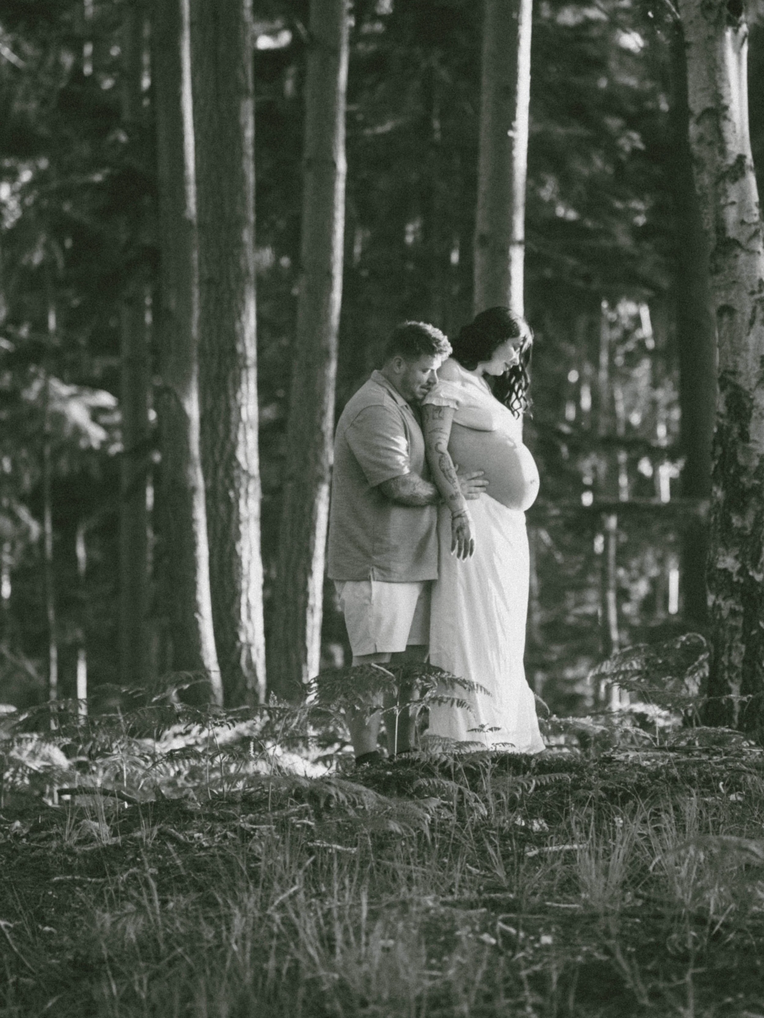 A black-and-white photograph of a man and a woman standing in a wooded area. The man has his head resting on the woman's belly, and they are both smiling. The woman is pregnant, wearing a flowing dress, and the man is wearing shorts and a shirt. They